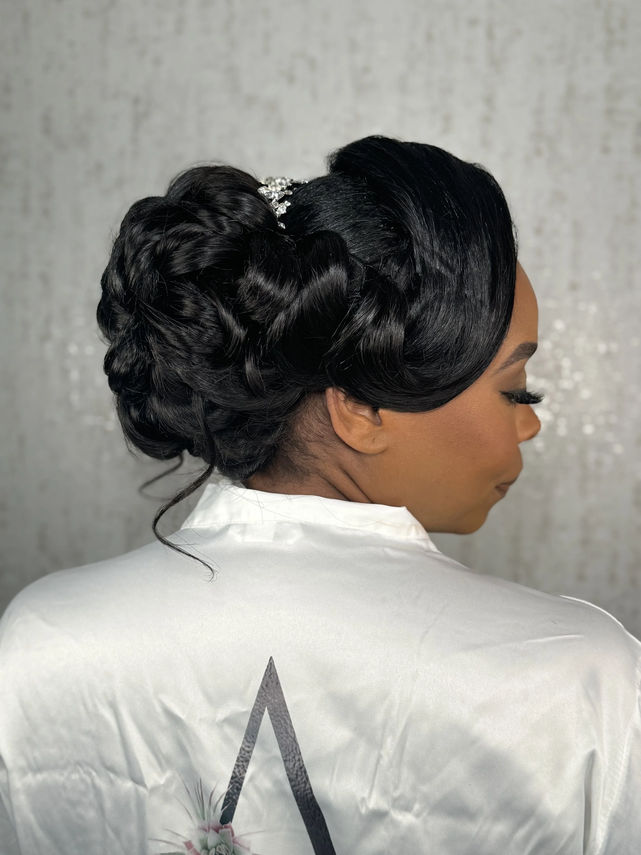 Side profile of a woman with elegant black updo hairstyle, adorned with a silver decorative hairpiece, wearing a white top, and standing against a light grey background.