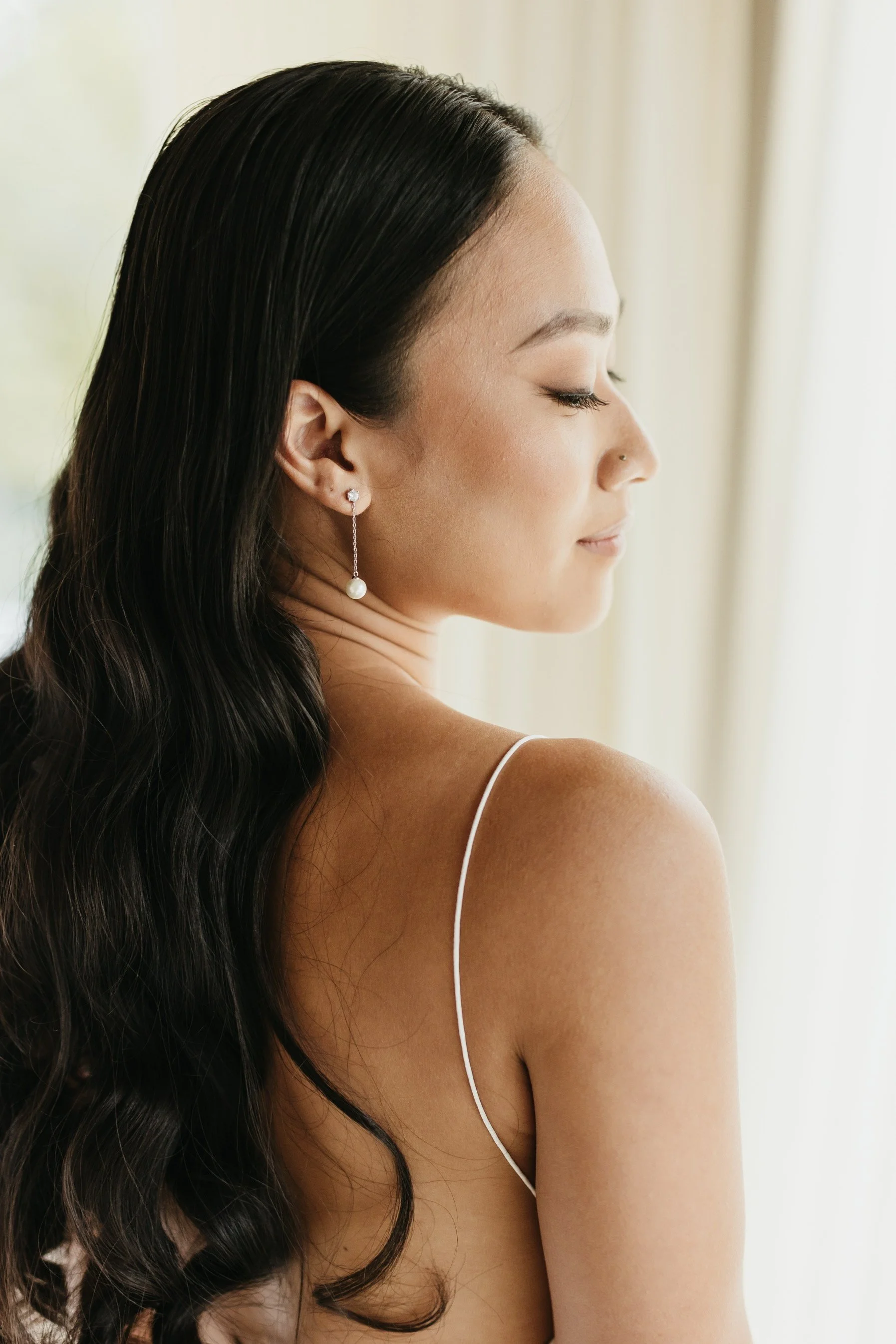 Asian Bride with long, dark hair wearing pearl earrings with chains, posing with her eyes closed in a softly lit room.