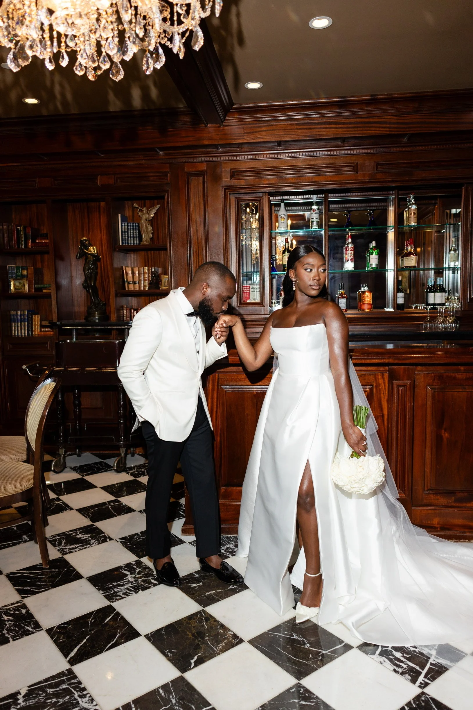 A bride and groom in a luxurious, wood-paneled room. The bride is wearing a white strapless wedding gown with a high slit, holding a bouquet of flowers and a small round purse. The groom is dressed in a white tuxedo jacket and black pants, leaning an