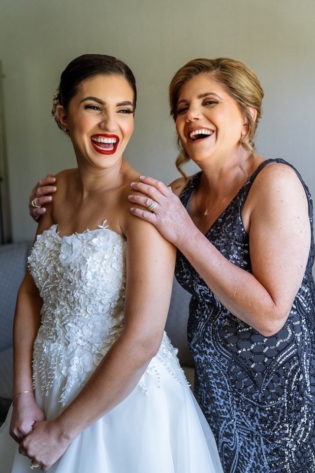 A bride and a woman, possibly her mother, laughing and smiling together, with the woman touching the bride's shoulder.
