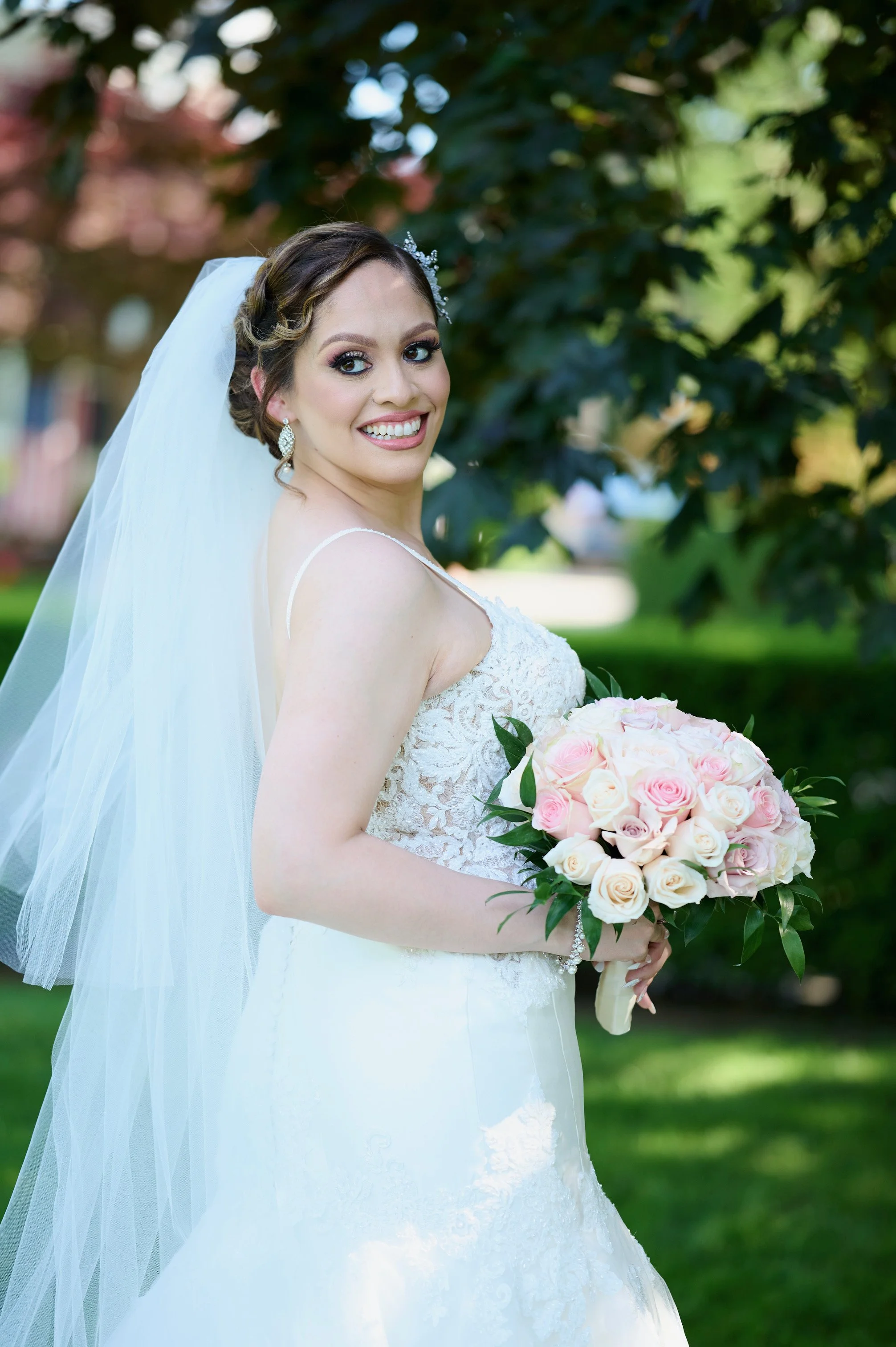 Brunette bride in a white wedding dress holding a bouquet of pink and white roses, standing outdoors with greenery in the background.