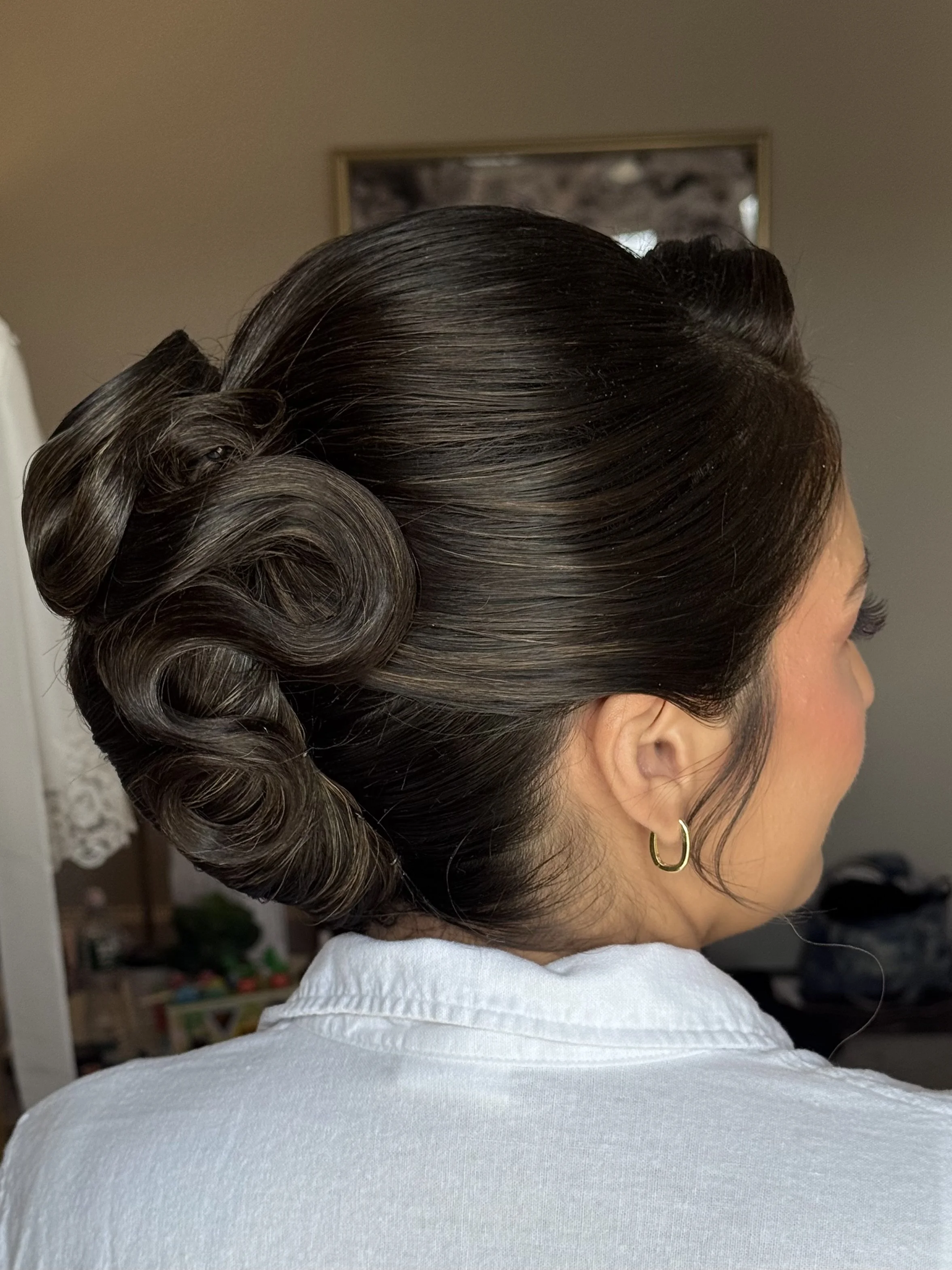 A woman's head with styled dark, shiny hair arranged in vintage curls, seen from the side, wearing a gold hoop earring and a white shirt.