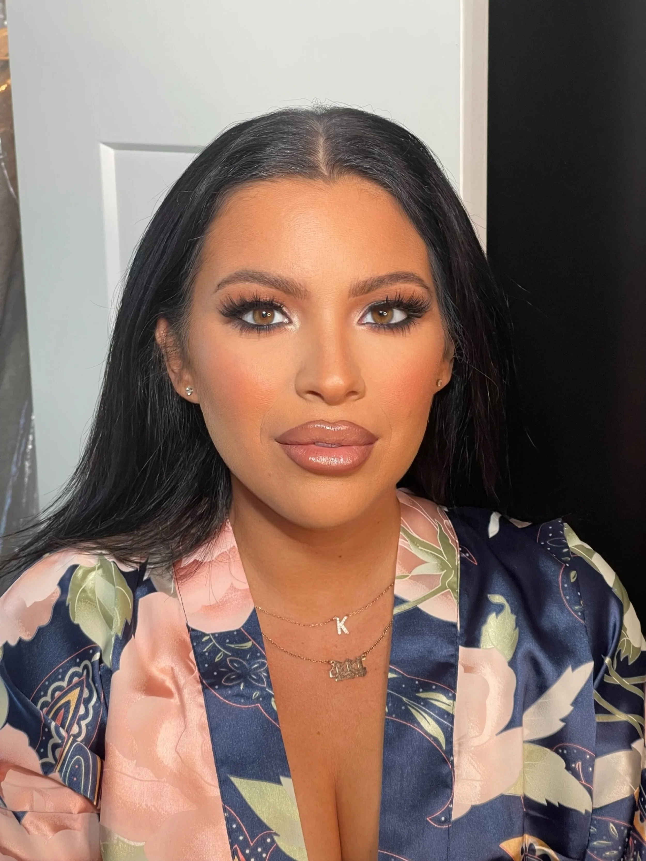 A woman with long black hair and tan skin, wearing makeup and jewelry, looking at the camera inside a room with a white door and dark background visible behind her.