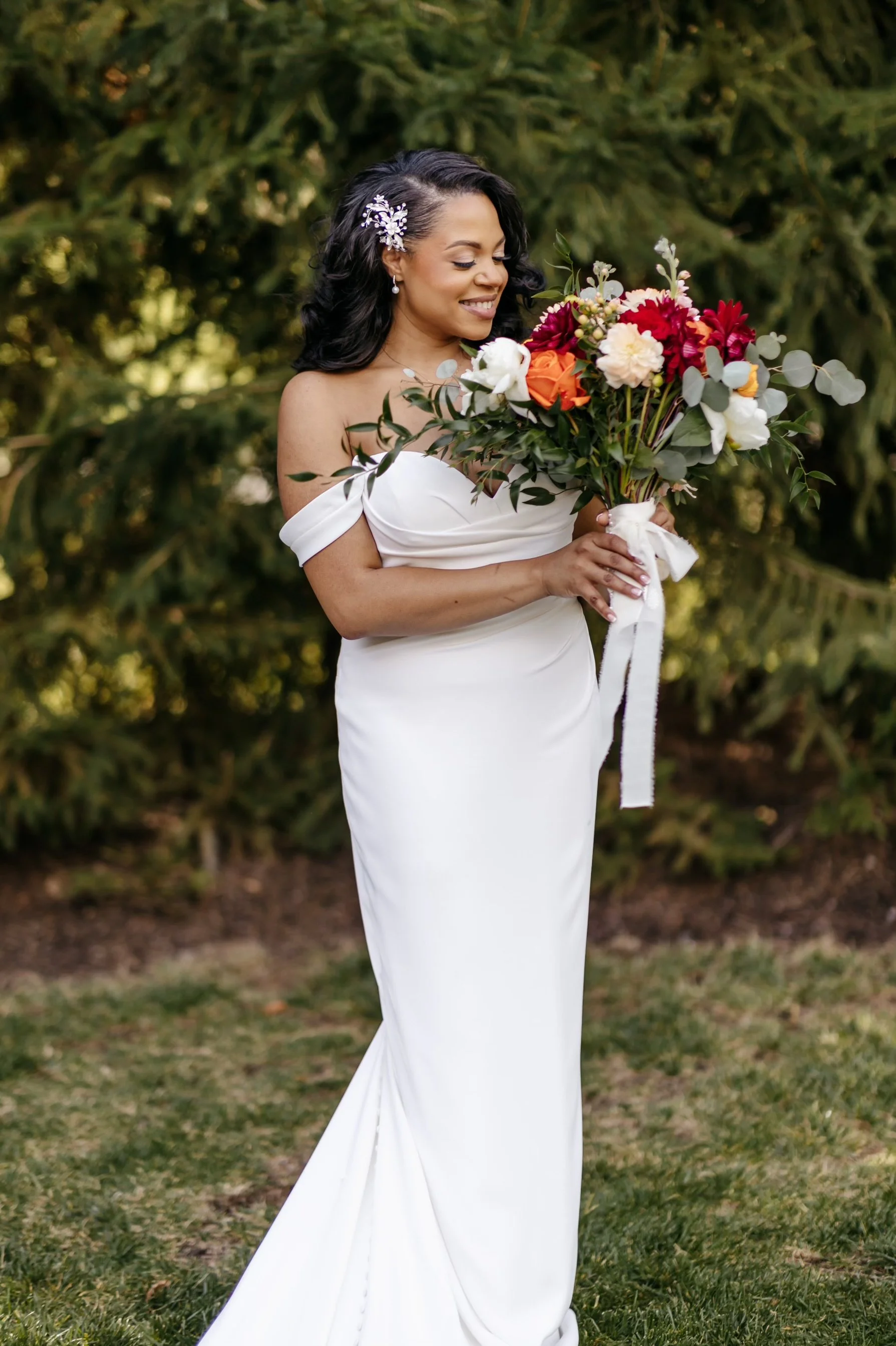 A woman in a white off-shoulder dress holding a bouquet of various colorful flowers, standing outdoors with trees and grass in the background.