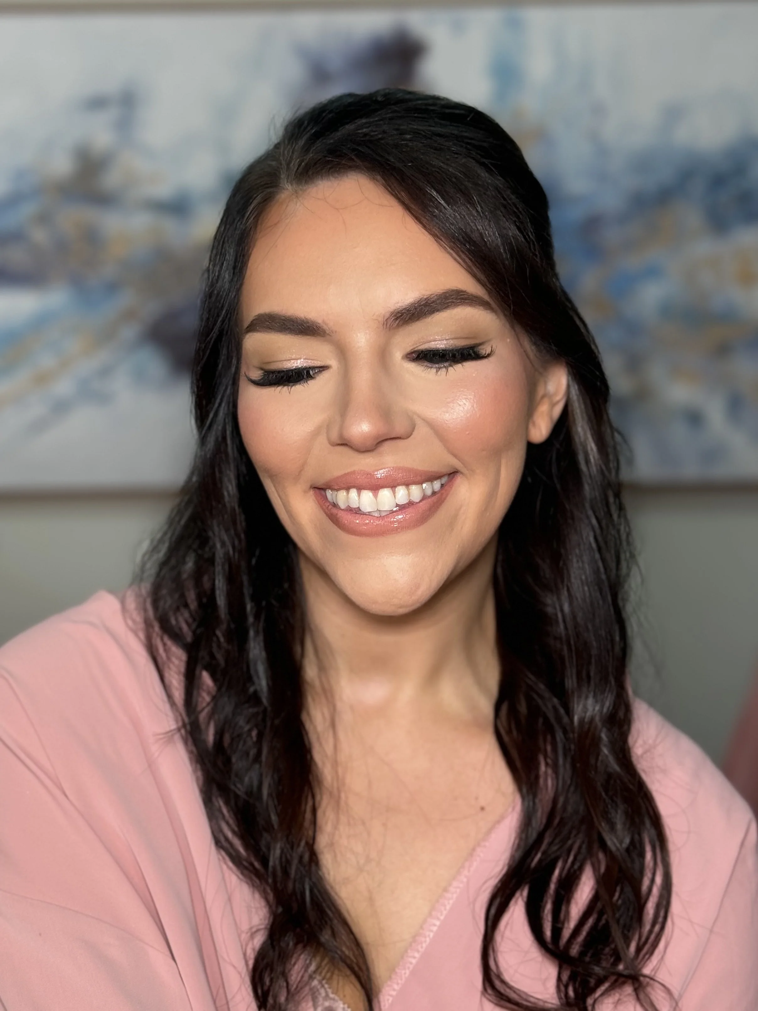 Close-up of a smiling woman with long dark hair, wearing a pink top, with a blurred abstract background.