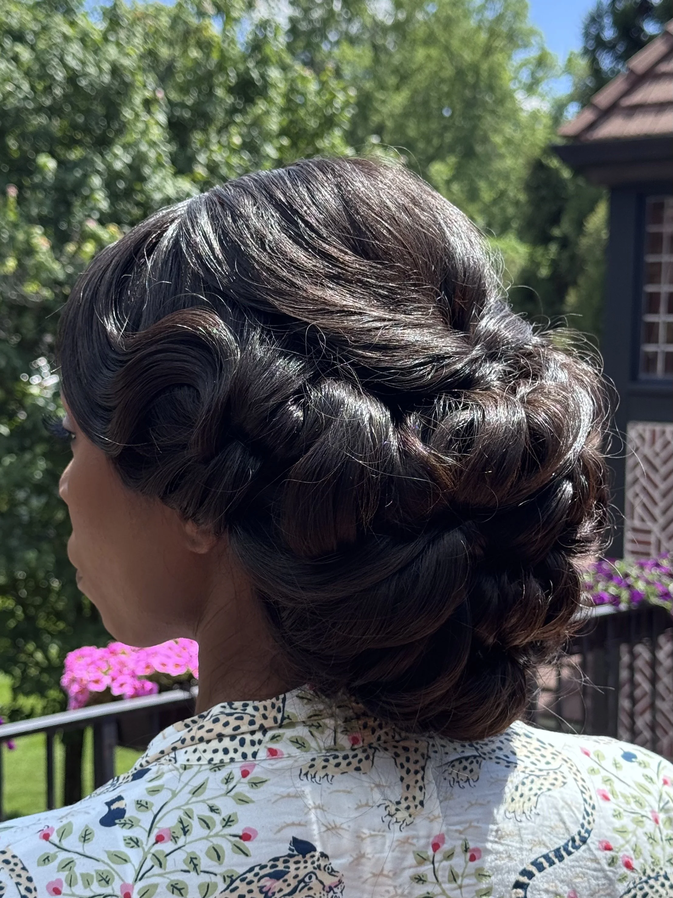 A woman with a vintage curled hairstyle, wearing a white shirt with a colorful floral pattern, standing outdoors with green trees and a house in the background.