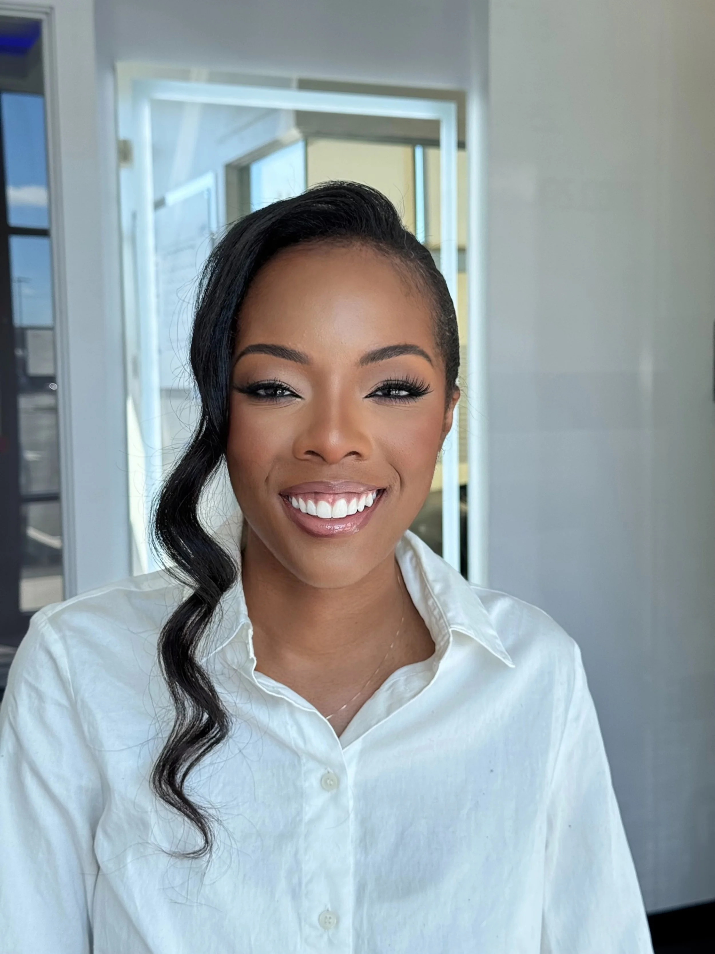 Close-up of a woman smiling, wearing a white button-up shirt, with a modern building and windows in the background.