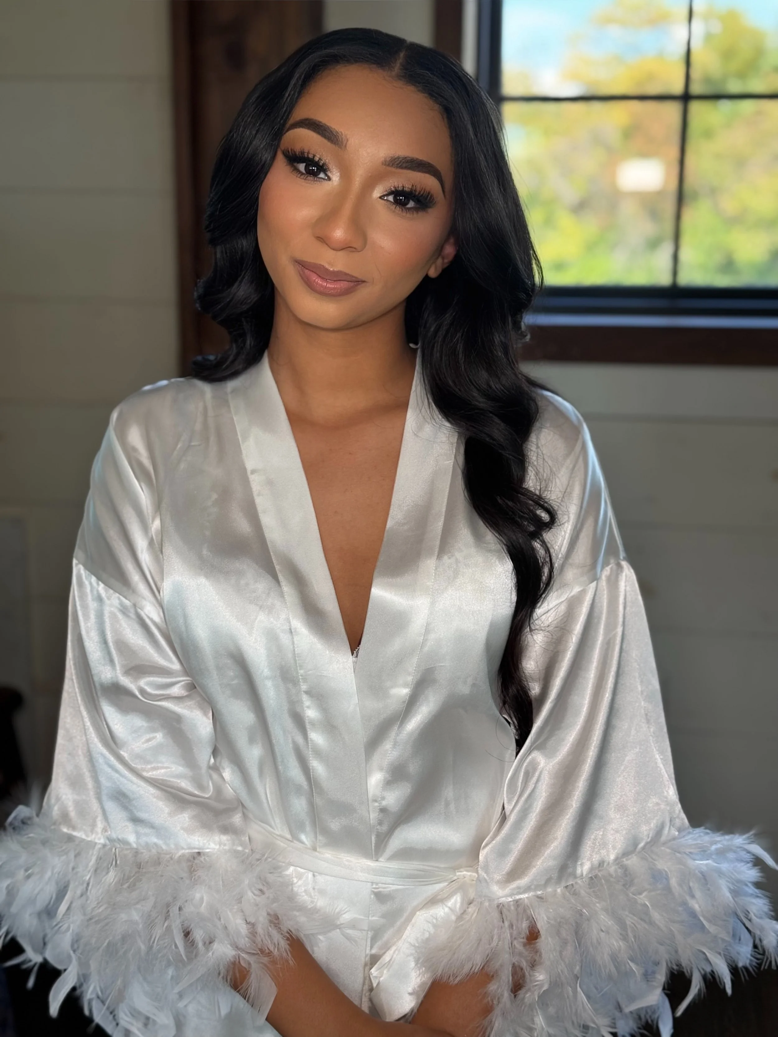A woman with long black hair and makeup, wearing a satin white robe with feathered cuffs, sitting indoors near a window with trees outside visible in the background.