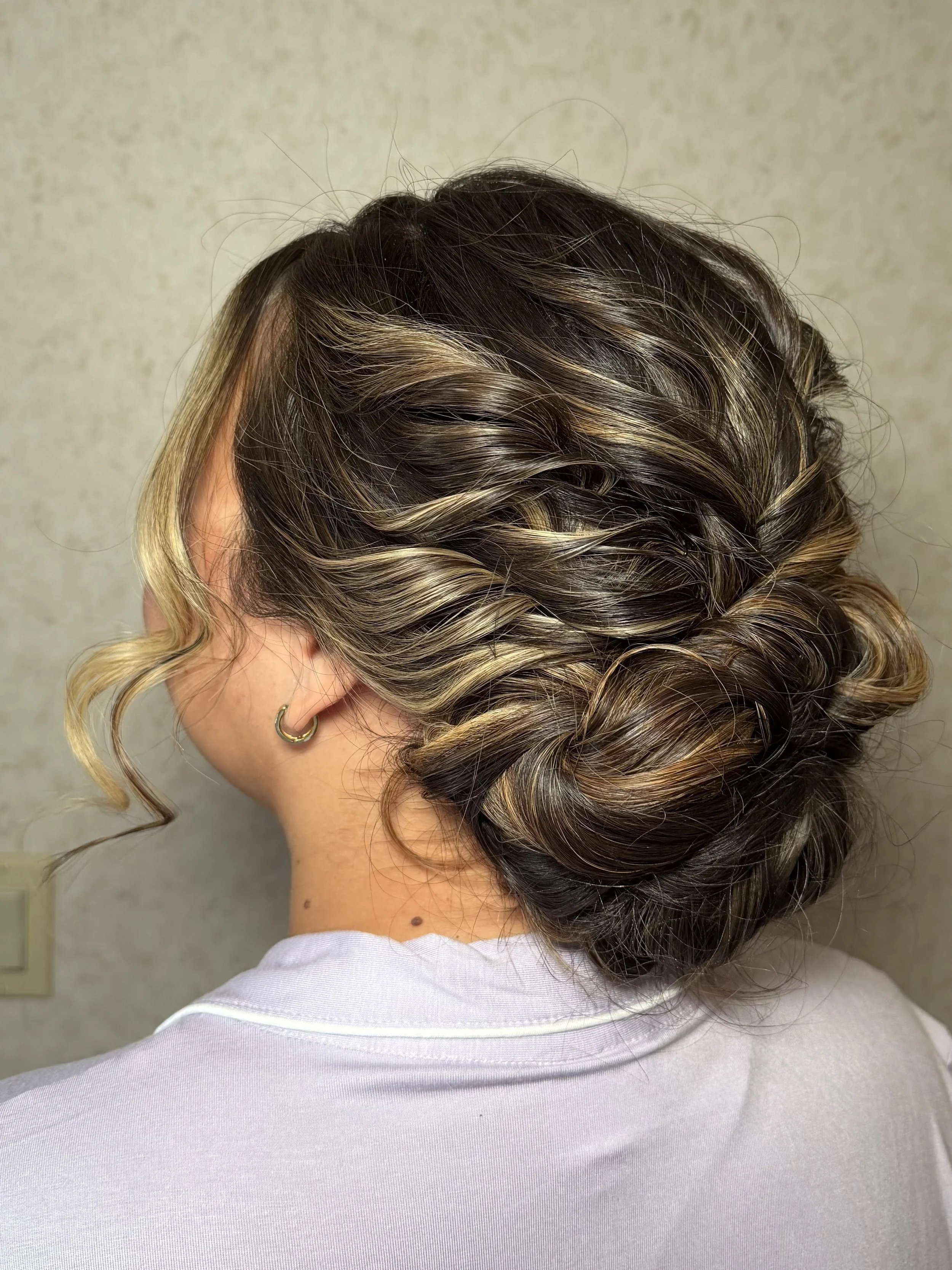 The image shows the back of a person's head with curly, styled hair featuring highlights, wearing a white shirt and a small hoop earring.