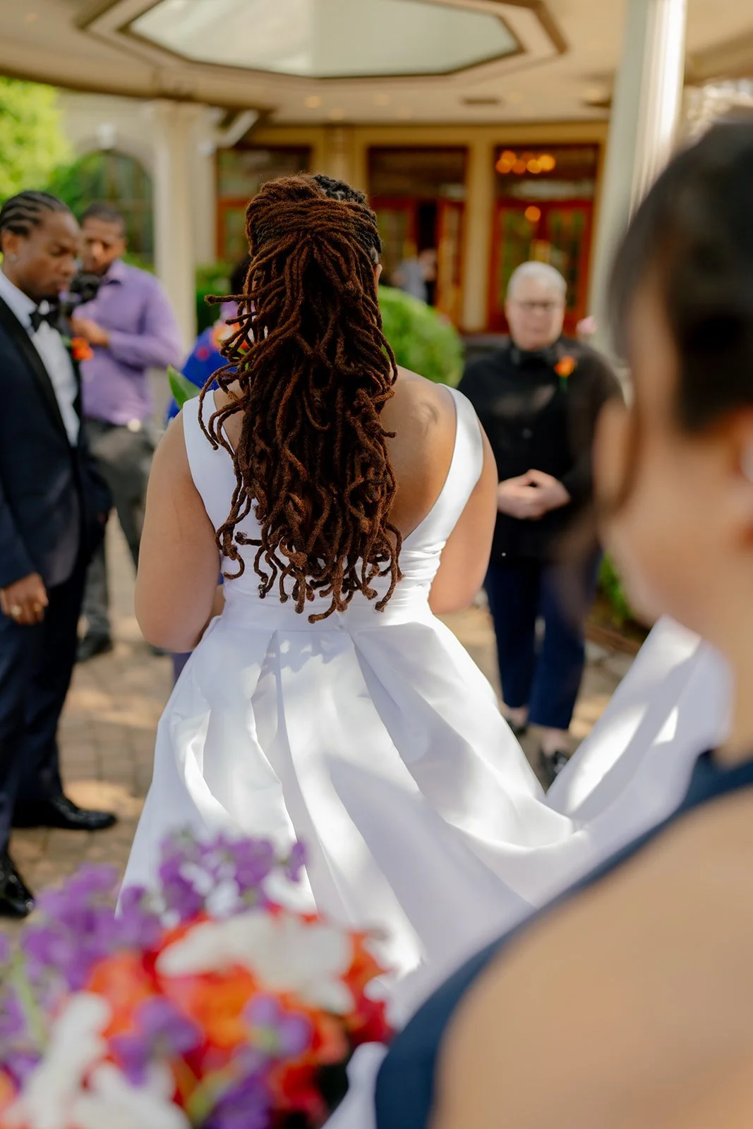 Back view of a woman in a white dress with long, curly dreadlocks at a wedding ceremony, with other guests in formal attire standing outdoors.