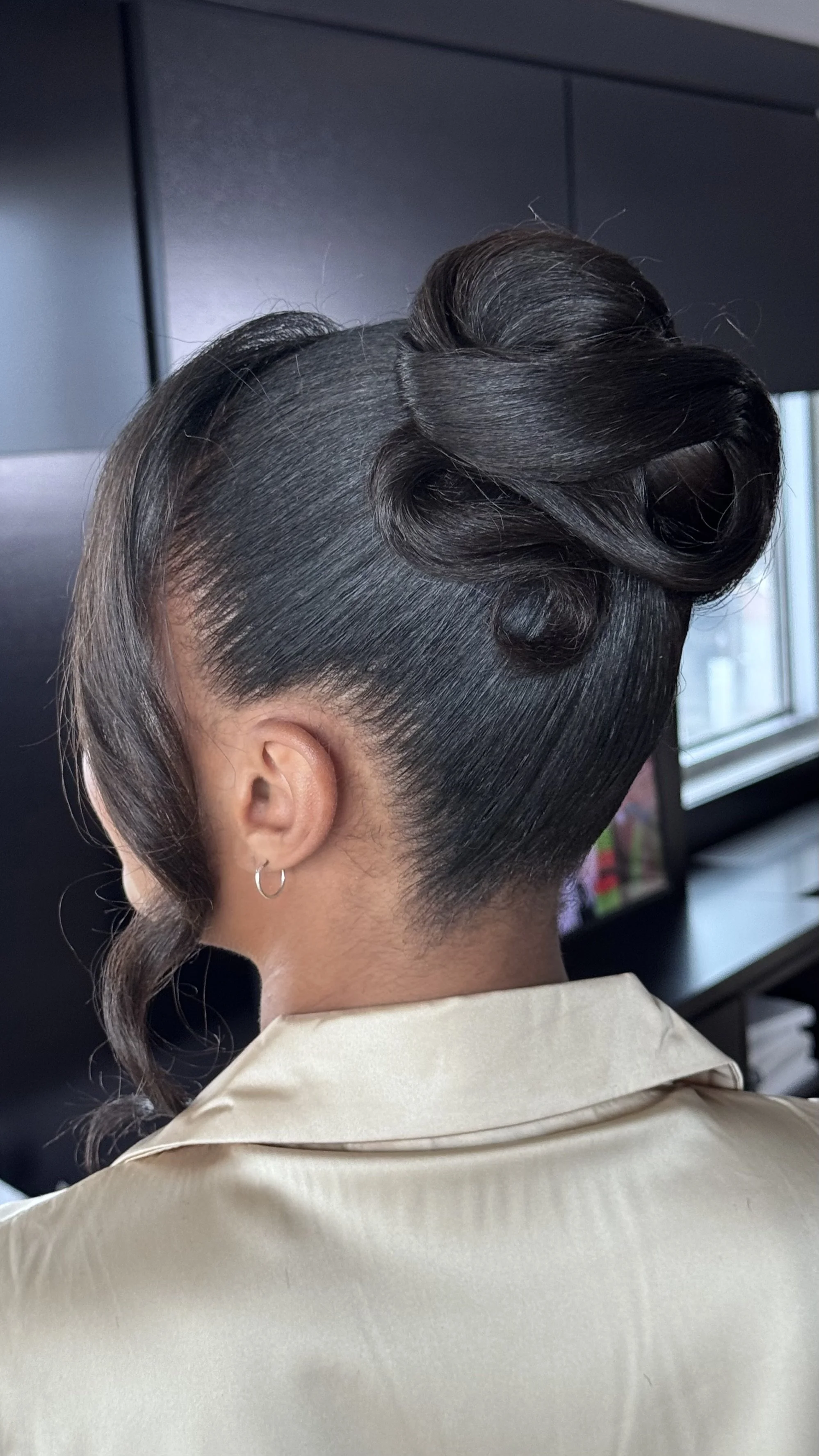 Close-up of a woman with an elegant updo hairstyle featuring a vintage-inspired roll and curled locks, wearing a light-colored satin jacket and a small hoop earring.