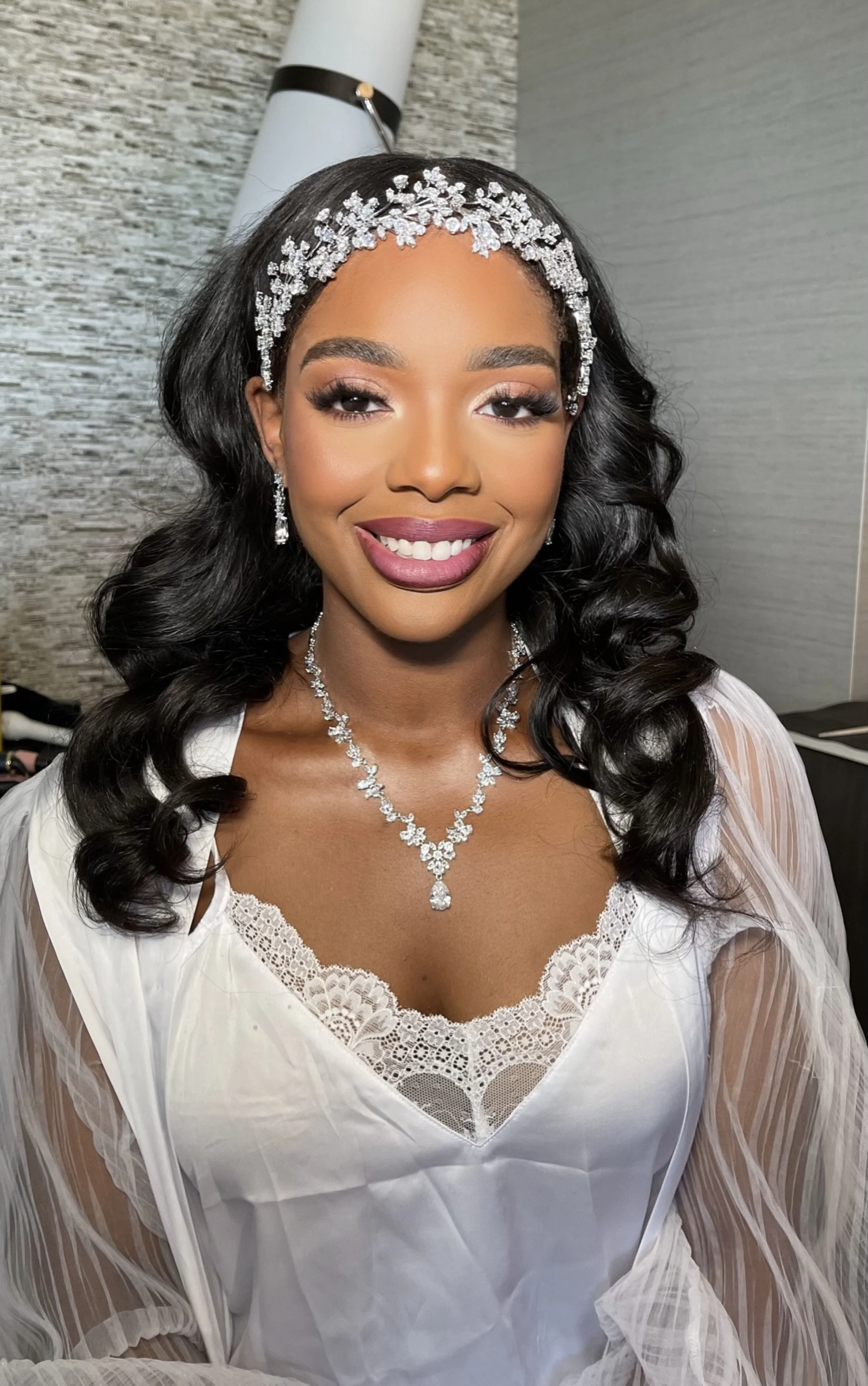 A woman dressed in white lace bridal attire, wearing a diamond necklace, earrings, and a tiara, smiling with dark curly hair styled in loose waves.