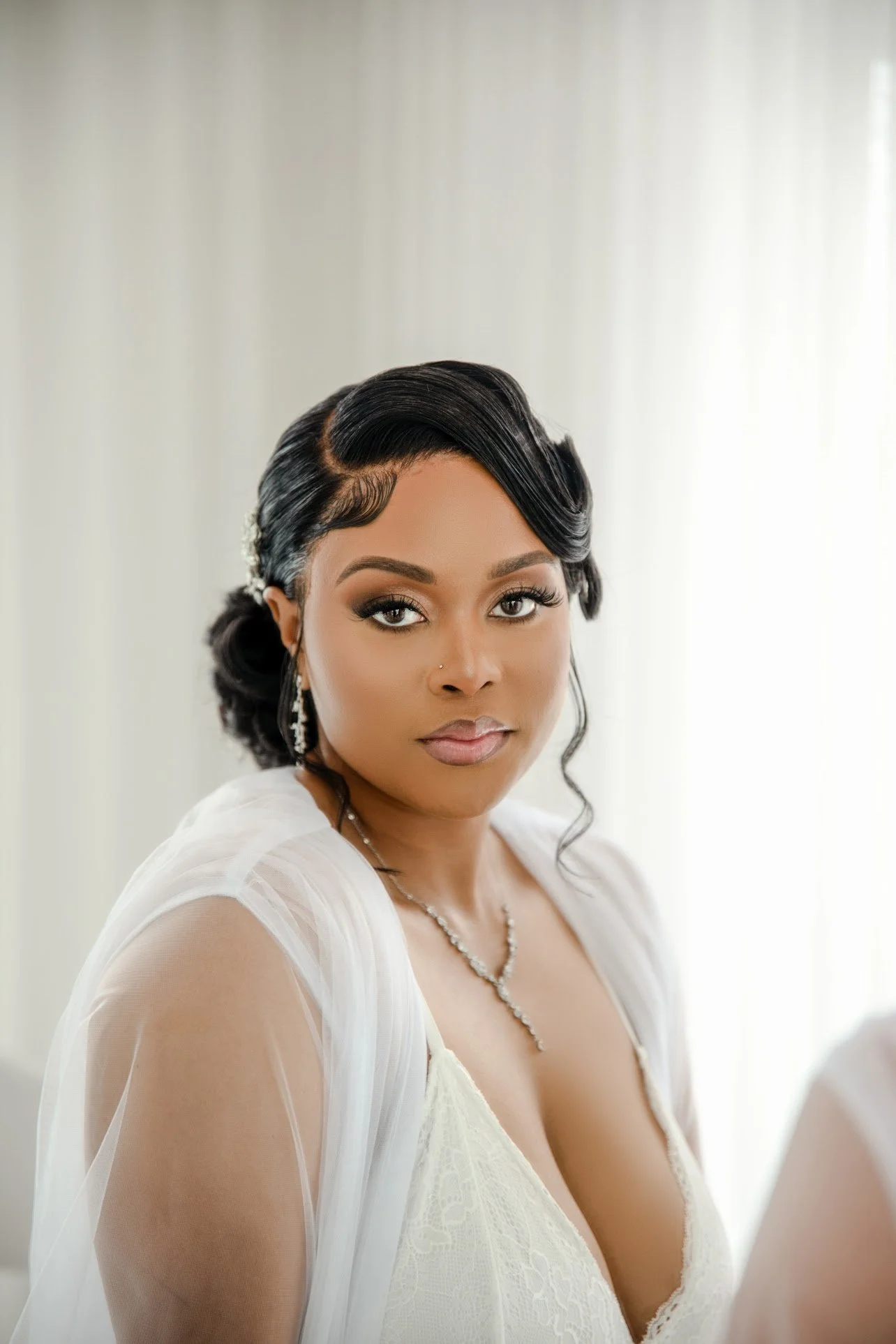 A portrait of a woman with dark hair styled in waves, wearing earrings and a white dress with sheer fabric on the shoulders. She has makeup with prominent eyelashes and lipstick, and is looking directly at the camera with a neutral expression.