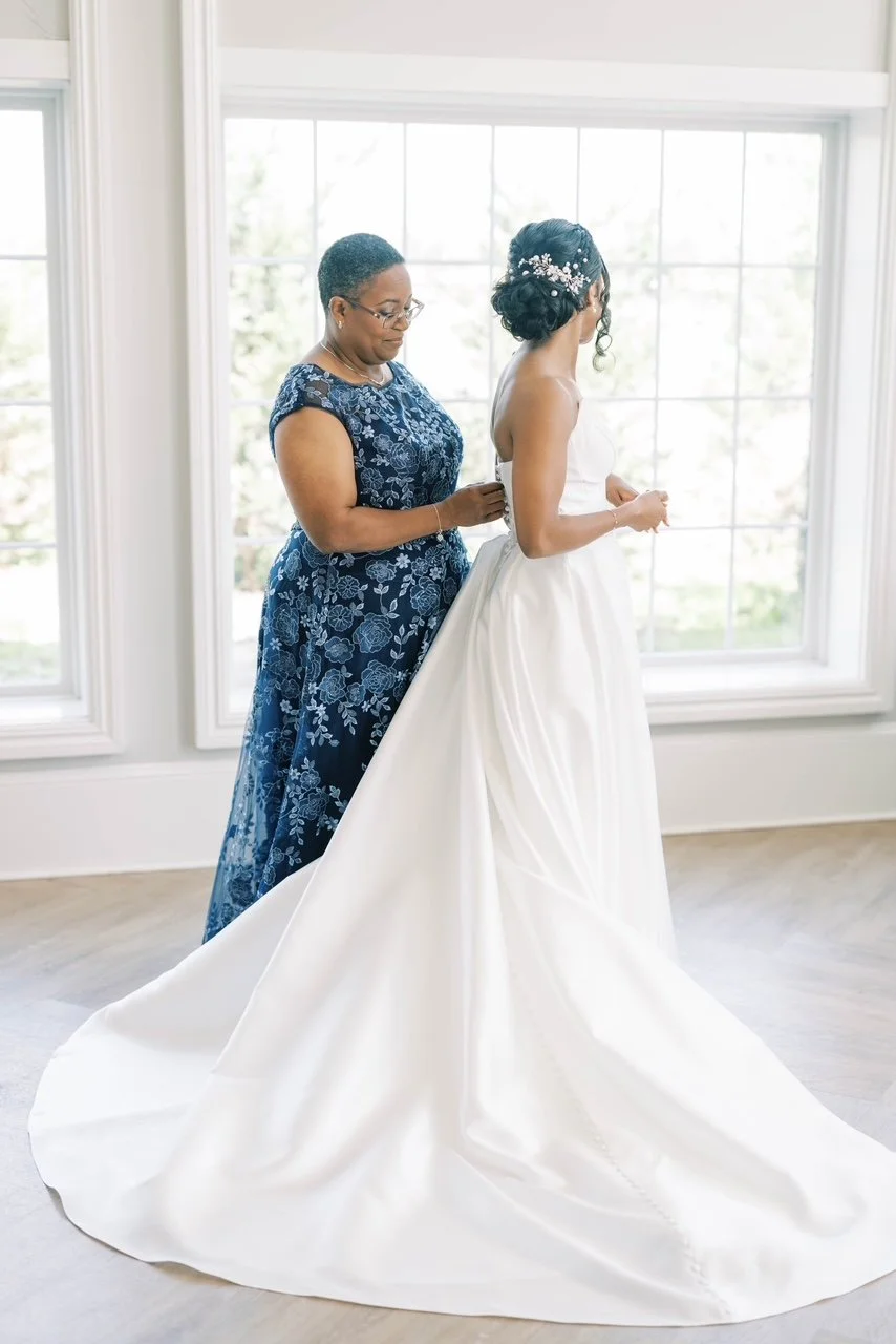 A bride getting ready in a white wedding gown, assisted by a woman in a blue floral dress, in front of large windows with natural light.