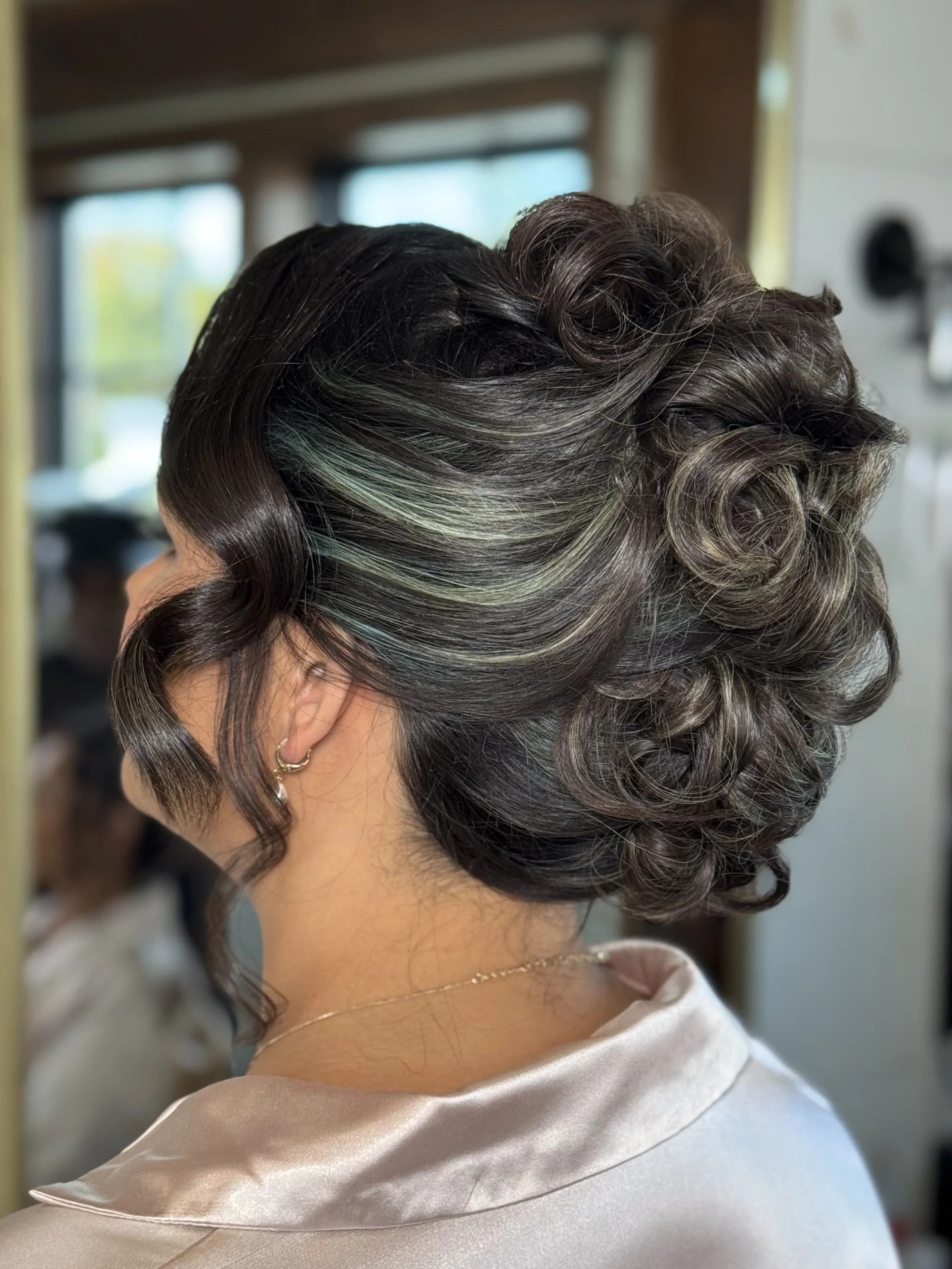 Close-up of a woman's elegant dark brown hair styled in intricate curls with subtle green highlights, wearing a shiny satin robe, in a room with a blurred background.