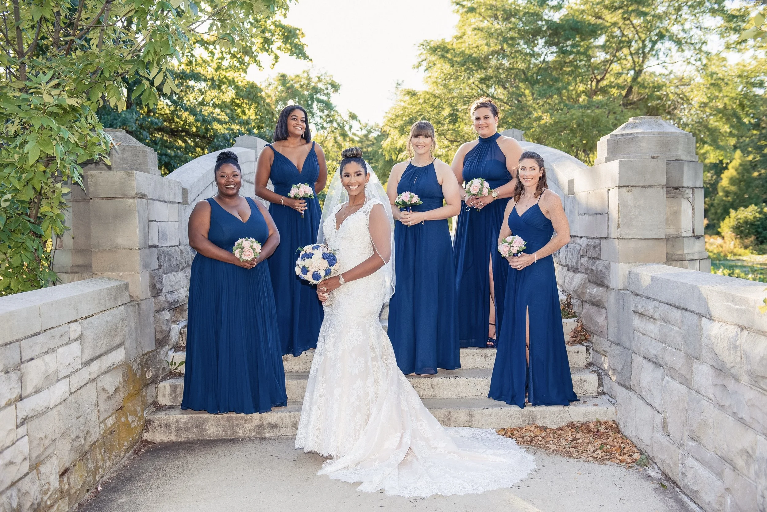 A group of seven women, including a bride, standing on steps outdoors. The women are dressed in dark blue bridesmaid dresses, and the bride is wearing a white wedding gown with a veil. They are holding pink and white bouquets and smiling, with green 