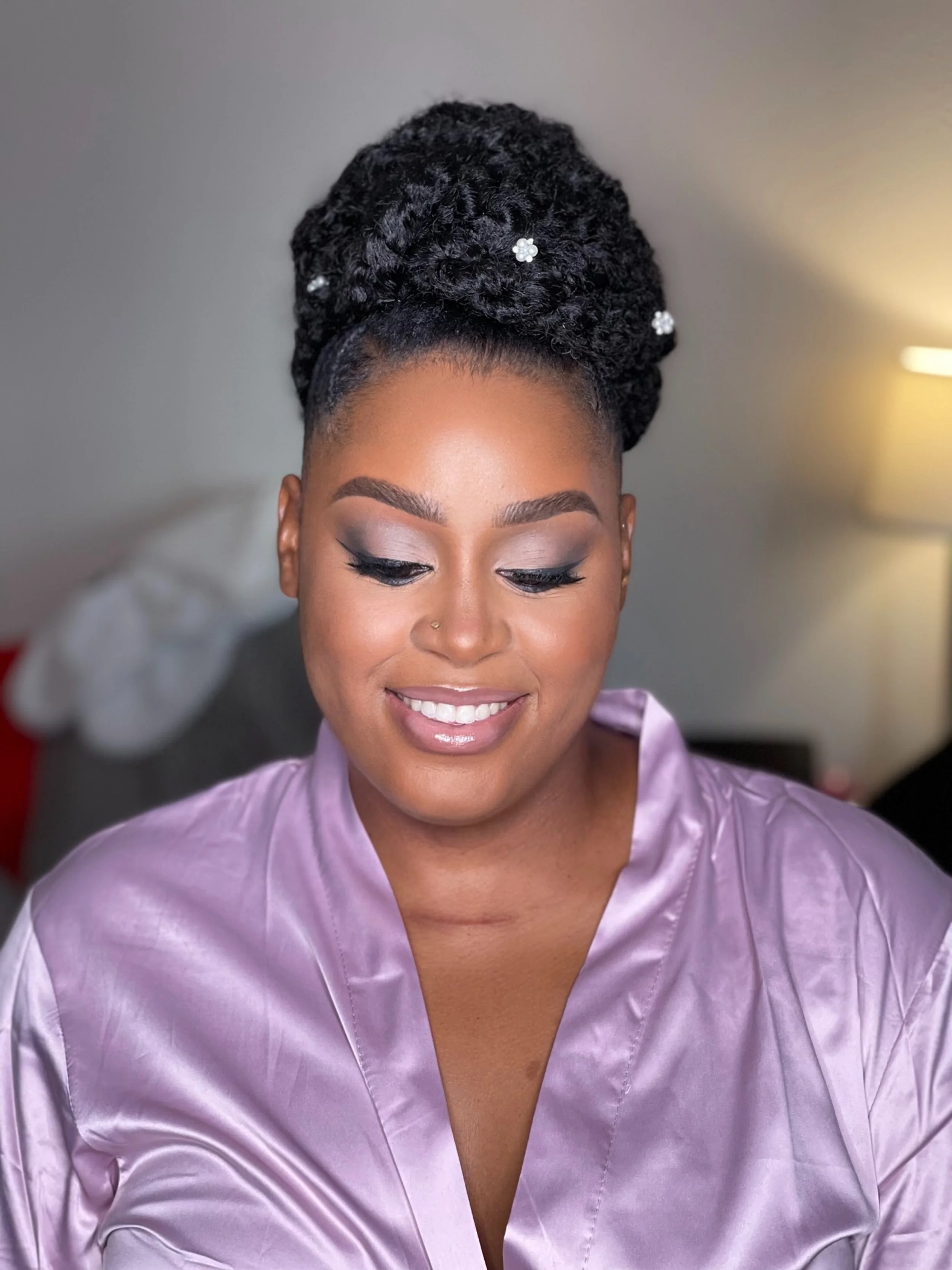 A woman with styled twists in her hair decorated with small pearl flower pins, wearing a light purple satin robe, smiling with makeup on her face.