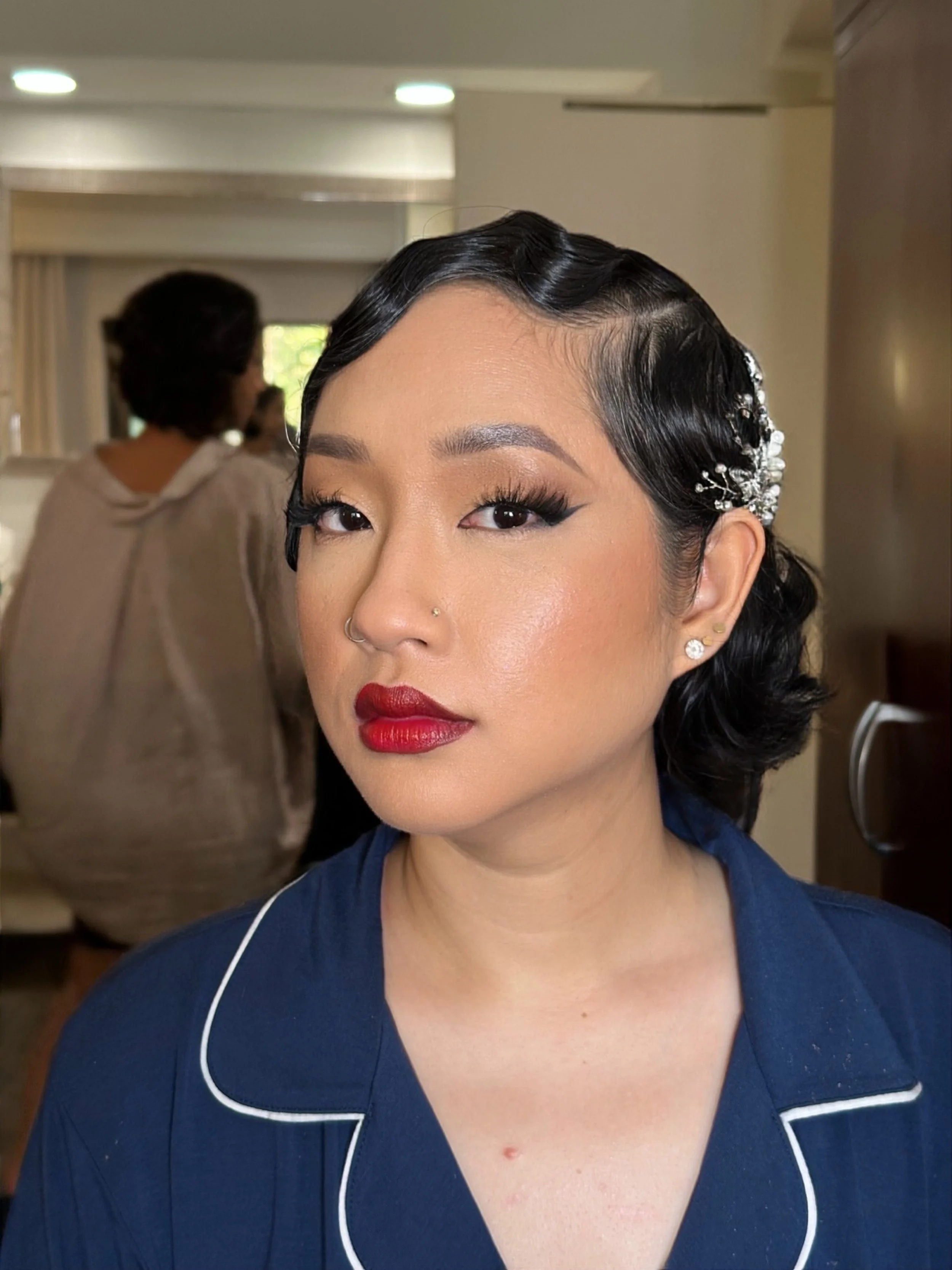 Close-up of a woman with vintage styled black hair, adorned with pearl hair accessories, wearing makeup with red lipstick, and dressed in a dark blue outfit with white piping.