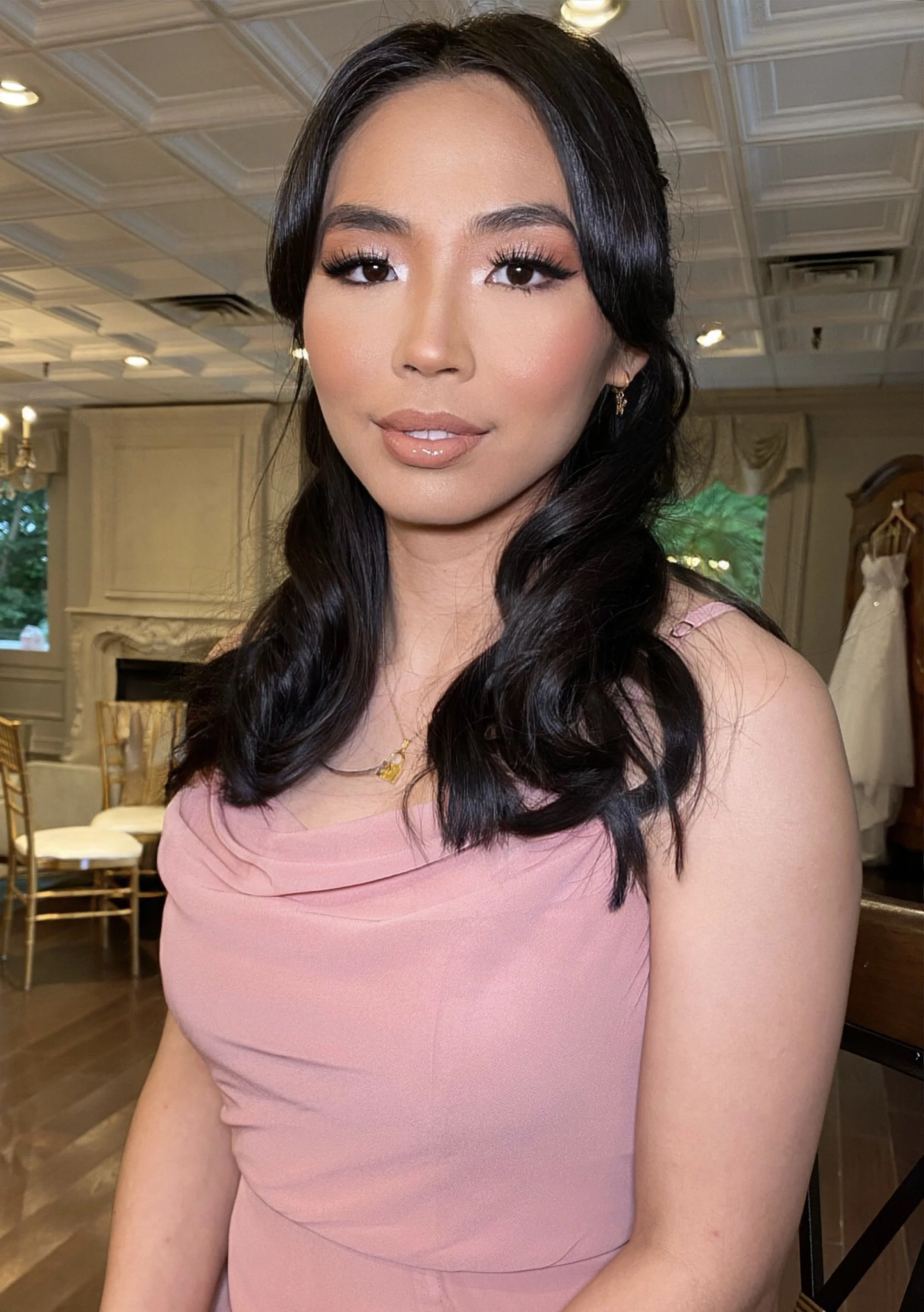 A woman with long dark wavy hair wearing a pink sleeveless dress, makeup, and earrings, sitting in a decorated room with a chandelier, chairs, a fireplace, and a wedding dress in the background.