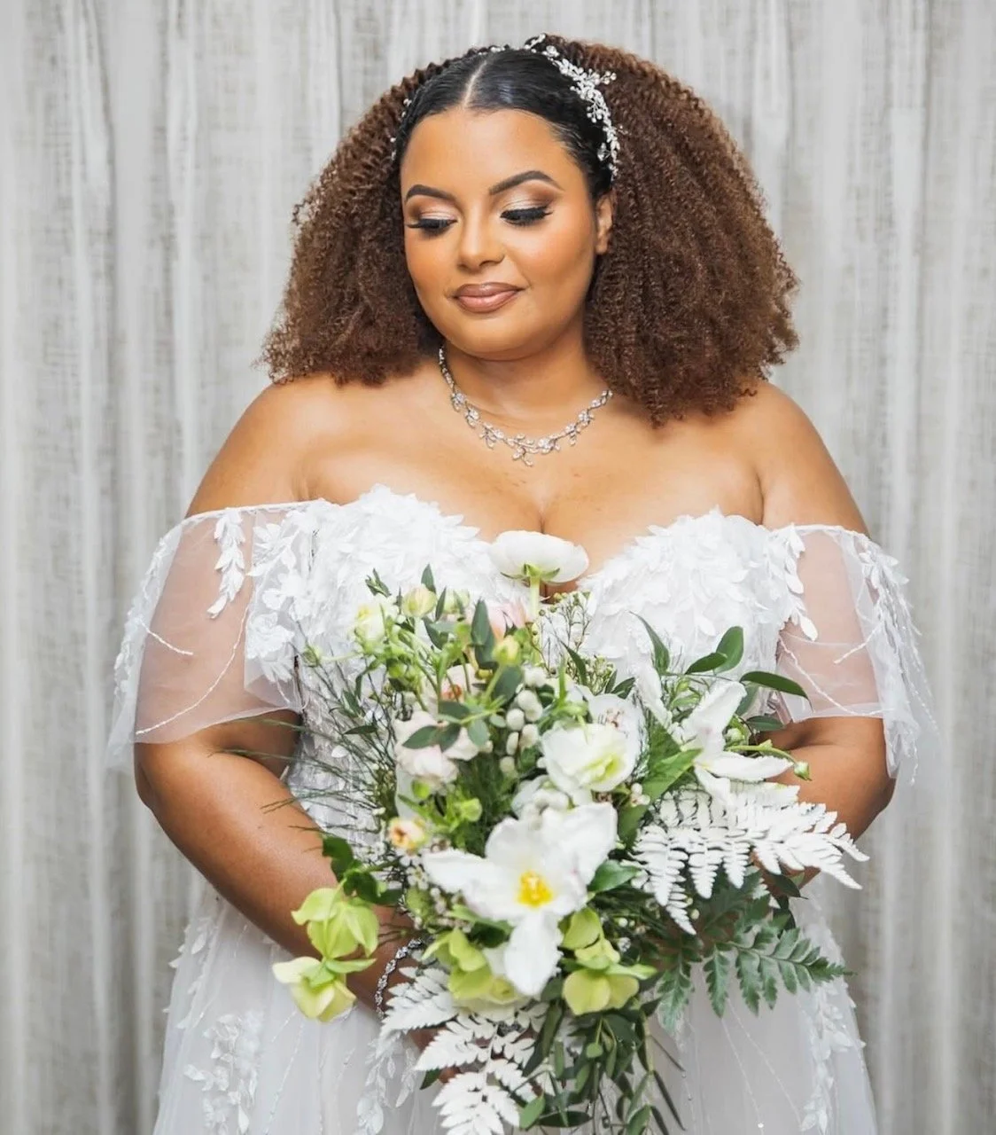 A bride in a white wedding dress holding a bouquet of white and green flowers, standing in front of a neutral background.