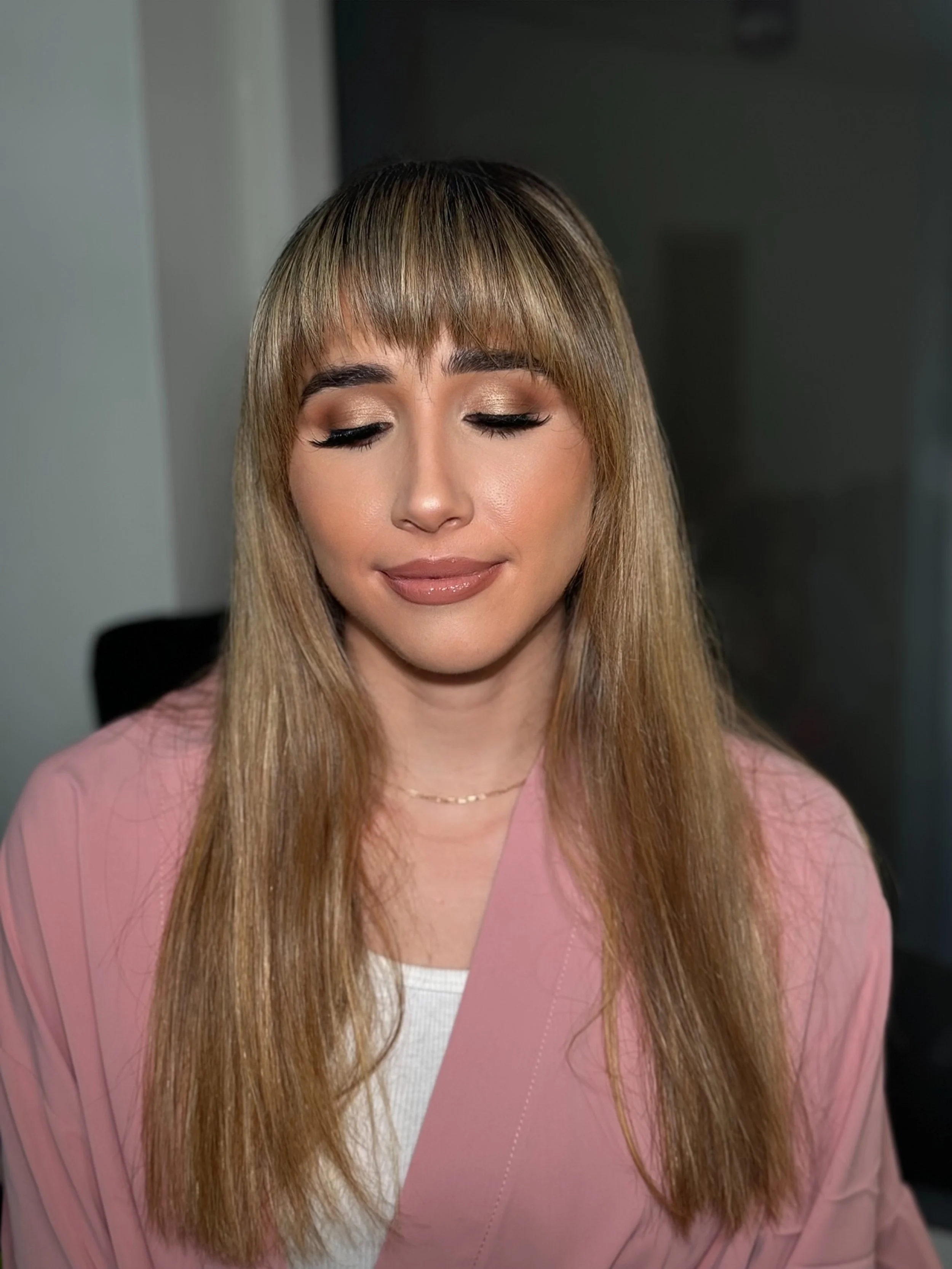 A woman with long, straight blonde hair and bangs, wearing a pink blazer and white top, with her eyes closed and a gentle expression.