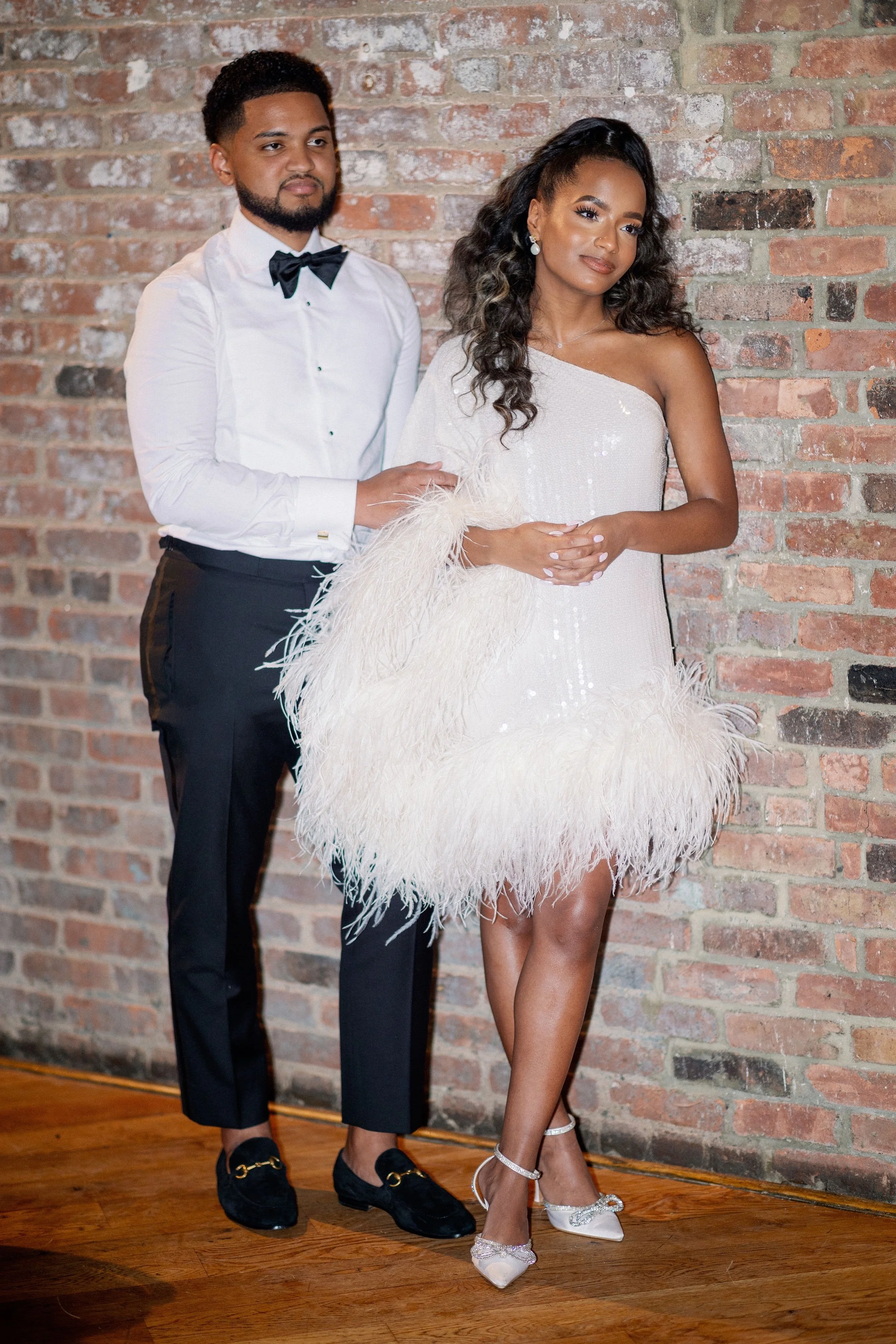 A man and woman dressed elegantly, standing against a brick wall. The man is in a white shirt with a black bow tie, black trousers, and black shoes. The woman is in a white, one-shoulder dress with feather details, wearing white high heels with ankle