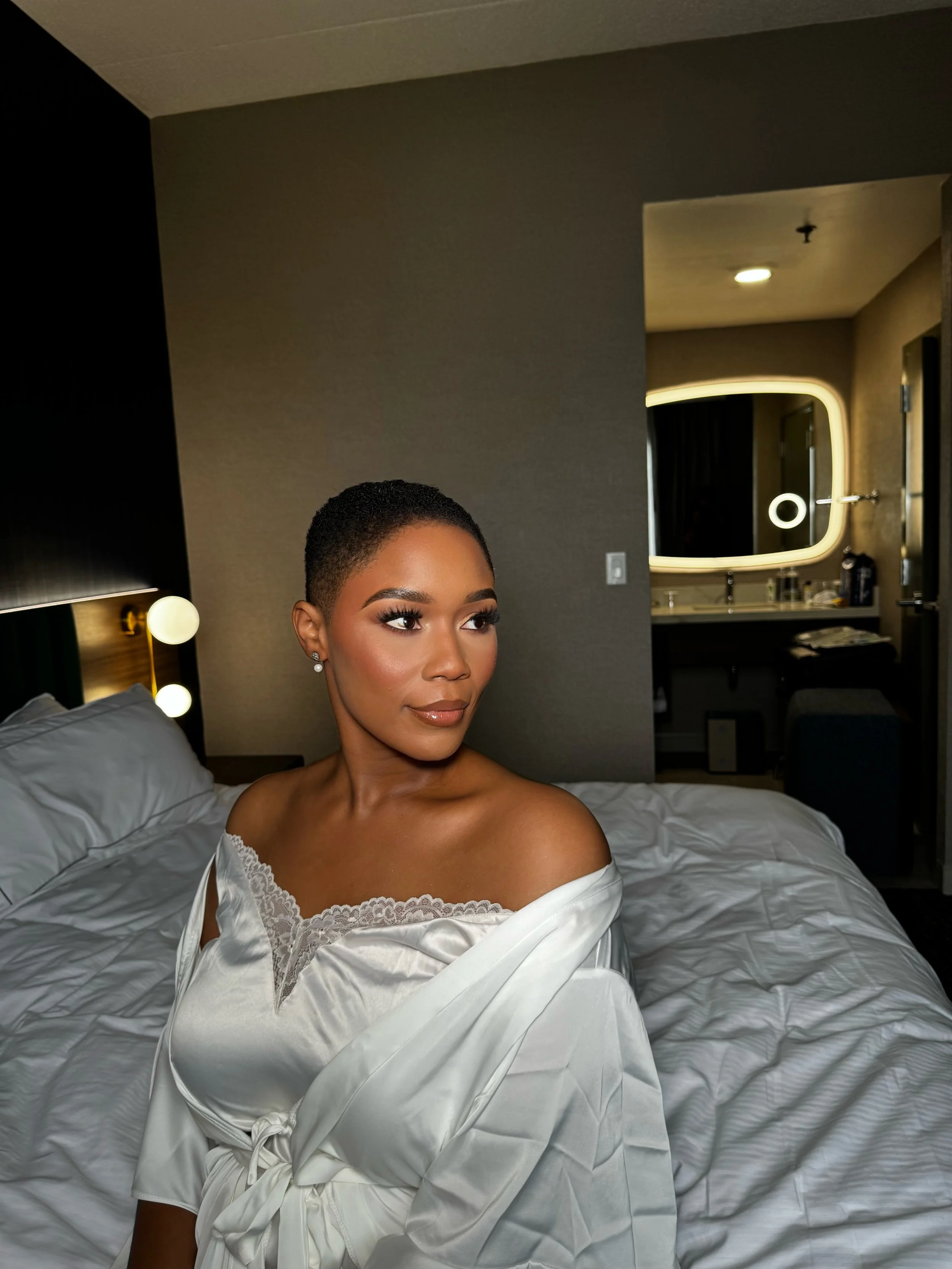 A woman with short black hair, wearing a white satin robe with lace details, sitting on a bed in a hotel room with a lit mirror and bathroom in the background.