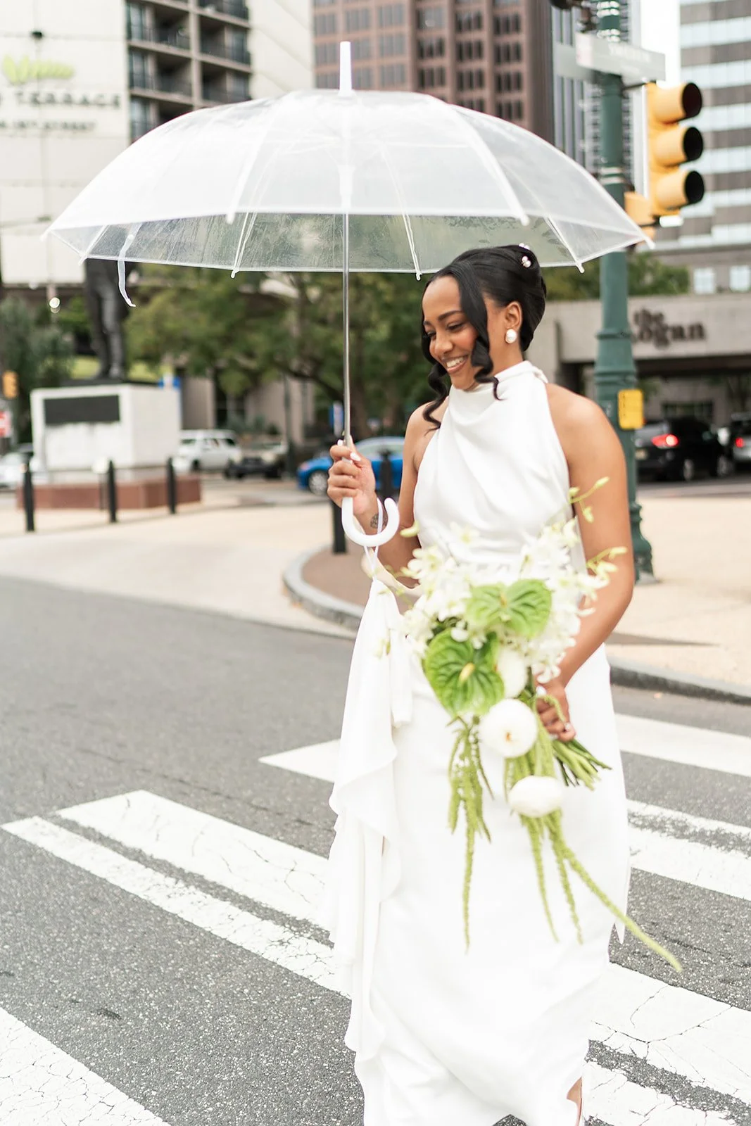 Woman in a white dress holding a bouquet of white flowers and an umbrella while walking on a city street.