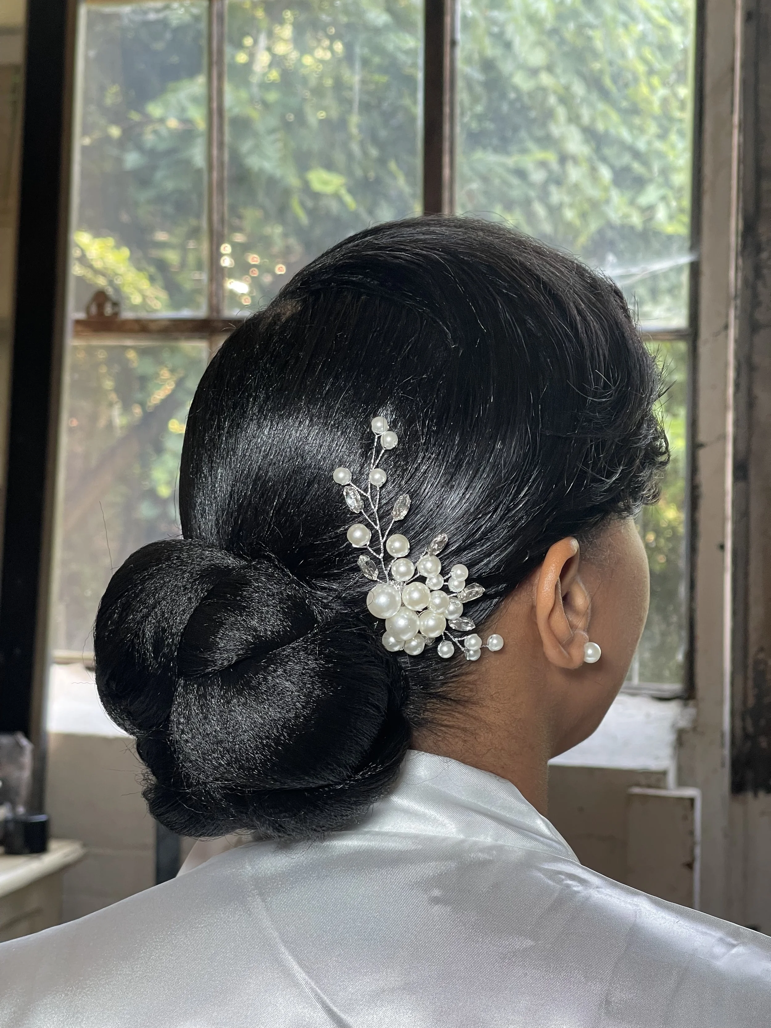A woman with black shiny hair styled in an elegant updo with a pearl and crystal hair accessory, wearing a white satin garment, standing indoors near a window with green foliage outside.