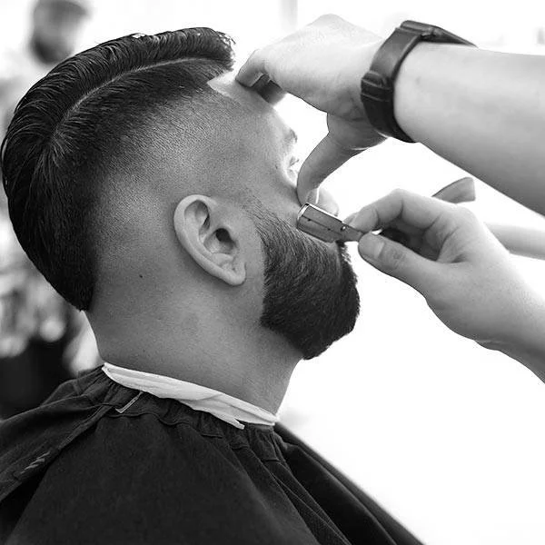 A man getting a haircut at a barber shop, with the barber shaving his beard using an electric razor.
