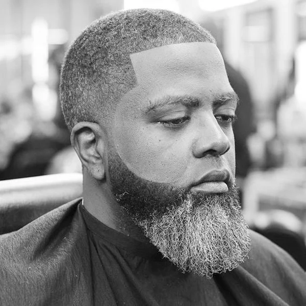 Man with a neatly trimmed beard and a fade haircut, sitting in a barber shop chair.