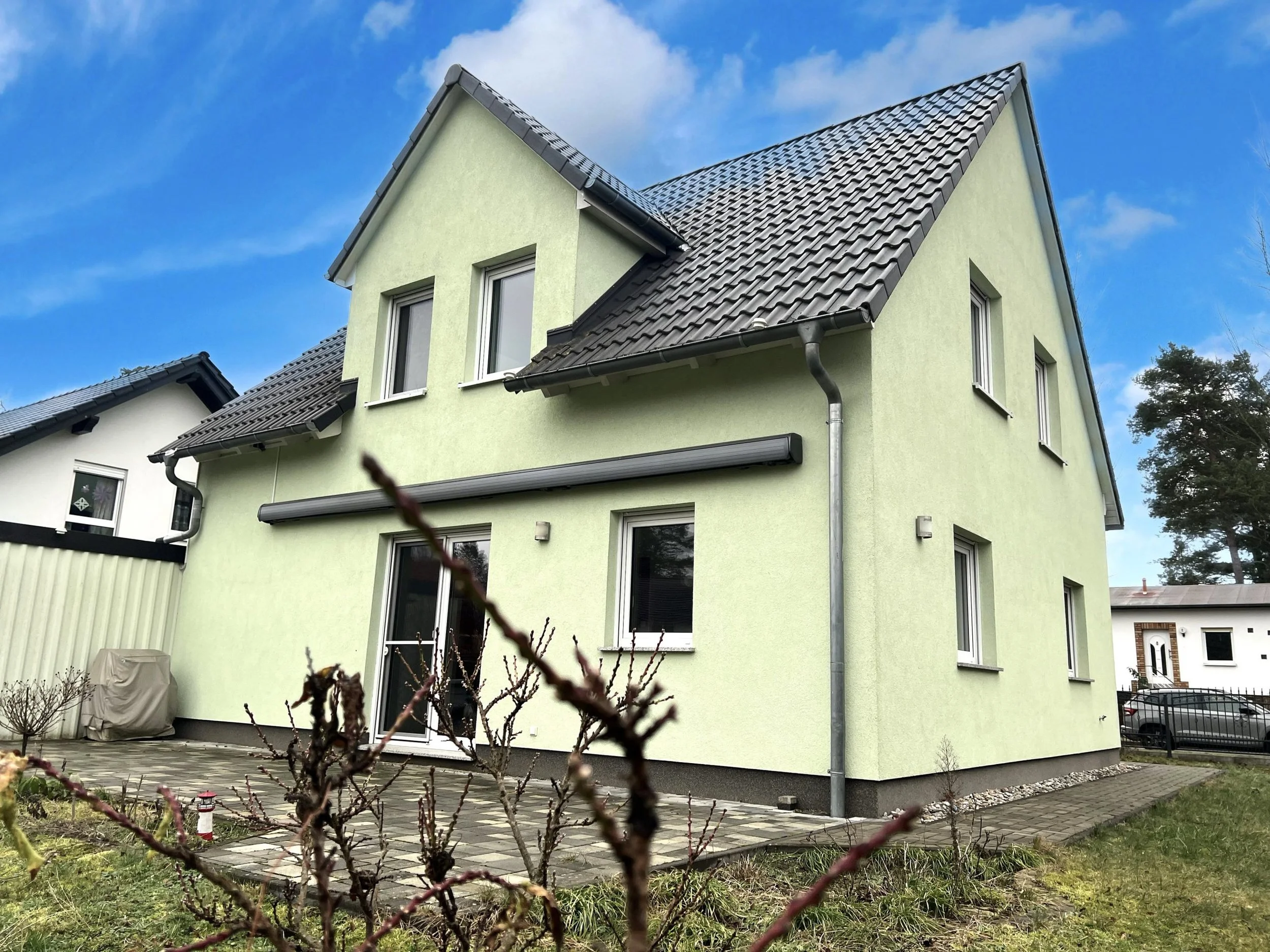 Ein grünes zweistöckiges Haus mit grauem Satteldach, weißen Fenstern und einer Terrasse mit Pflastersteinen, umgeben von einem Garten mit Sträuchern und einem Weg.
