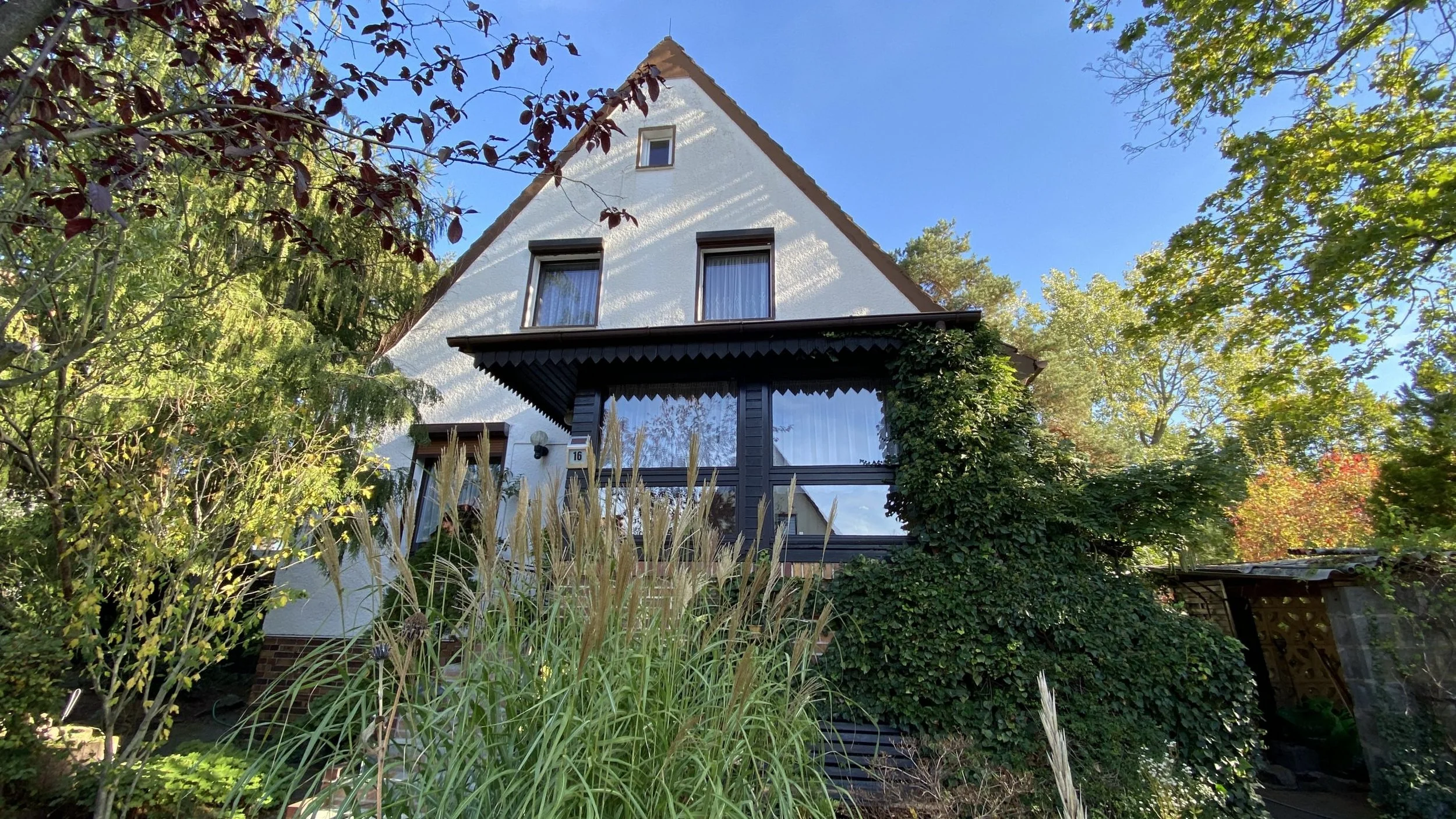 Ein Haus mit weißen Wänden und braunem Dach, umgeben von Bäumen und Sträuchern, ein Blumentopf an der Hauswand, klare blauer Himmel.