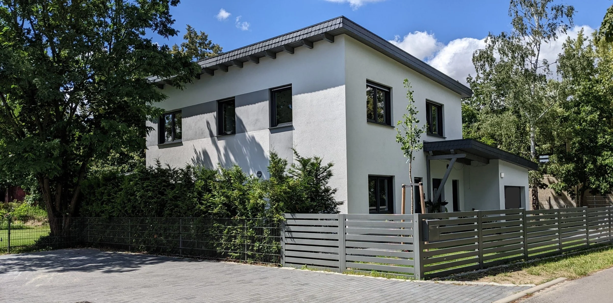 Modernes zweistöckiges Haus mit weißer Fassade, dunklen Fenstern und Flachdach, umgeben von Bäumen und einem Garten, eingezäunt mit einem grauen Zaun.