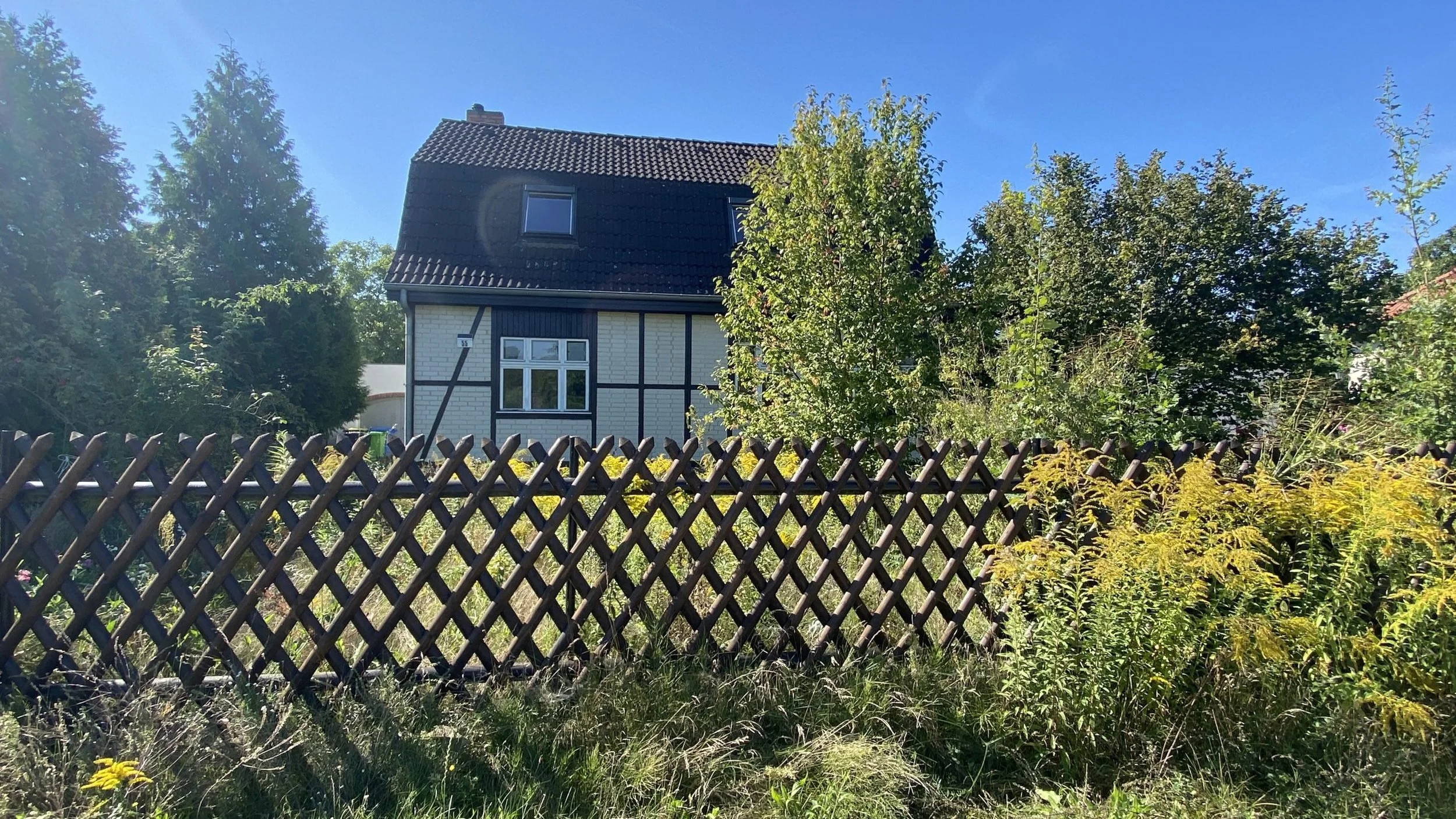 Ein Haus mit weißer Fassade und dunklen Holzstreben, umgeben von Bäumen, auf einem Zaun aus Holzlatten, bei sonnigem Himmel.