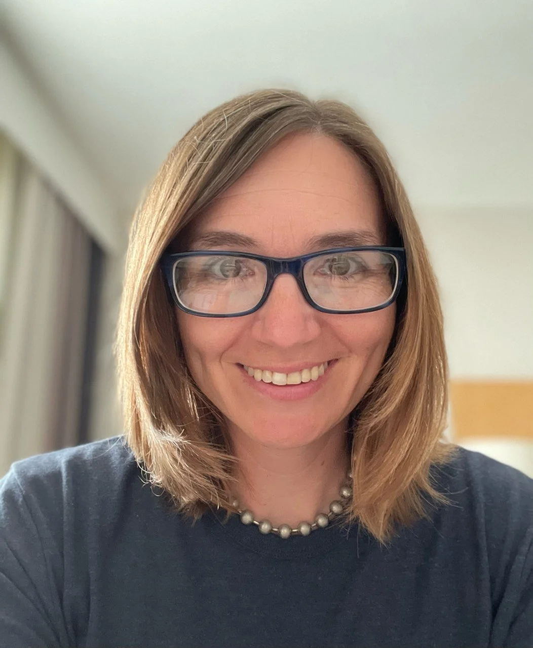 Headshot of Bettie, a white woman with chin length straight blonde hair and glasses, smiling at the camera