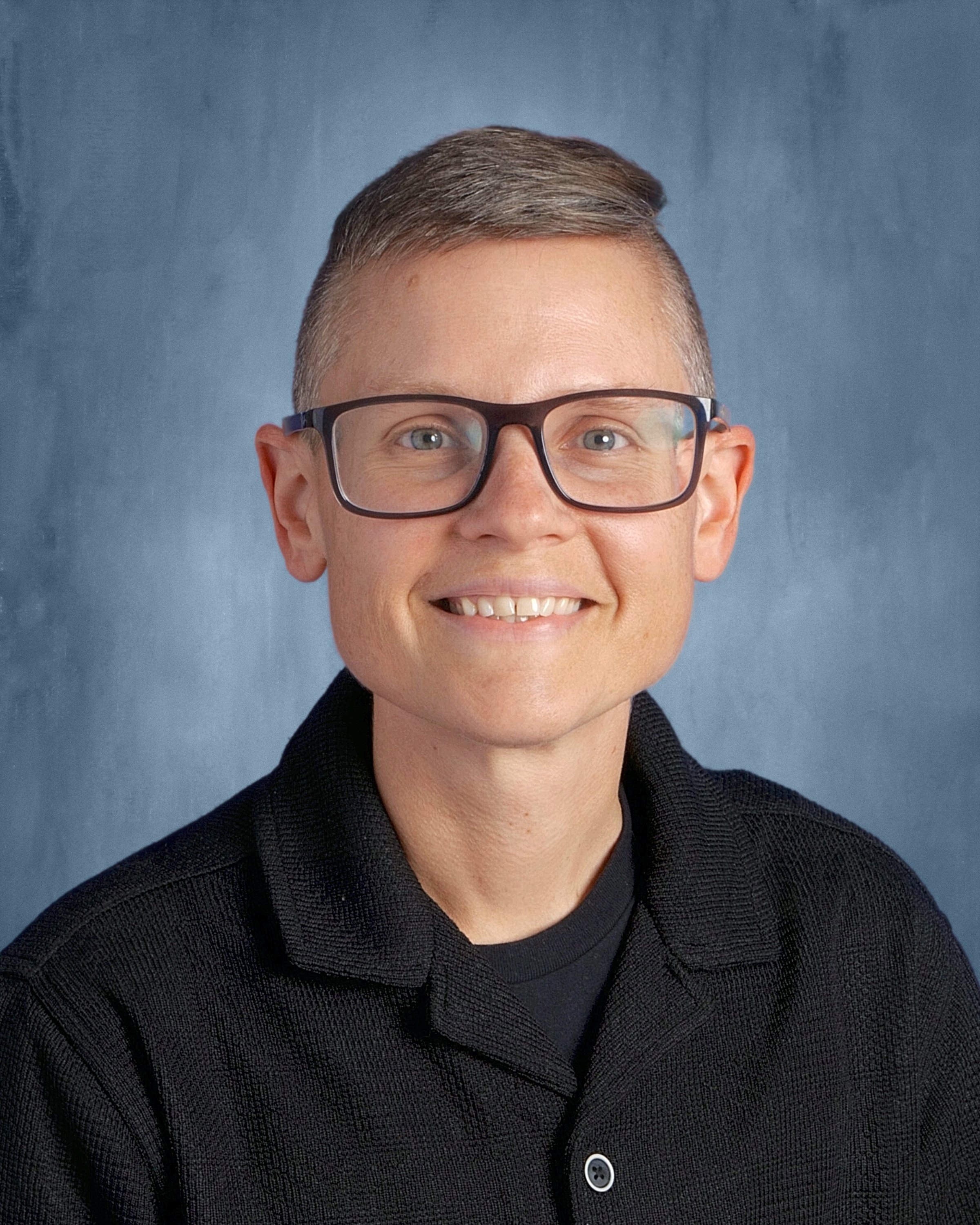 Morgan is a white person with very short hair and black glasses, smiling against a gray-blue background