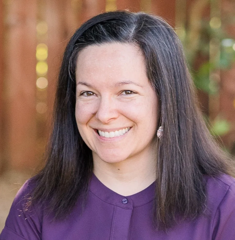 Leah is a white woman with straight brown hair parted at the side. She is grinning at the camera in front of a brown fence.