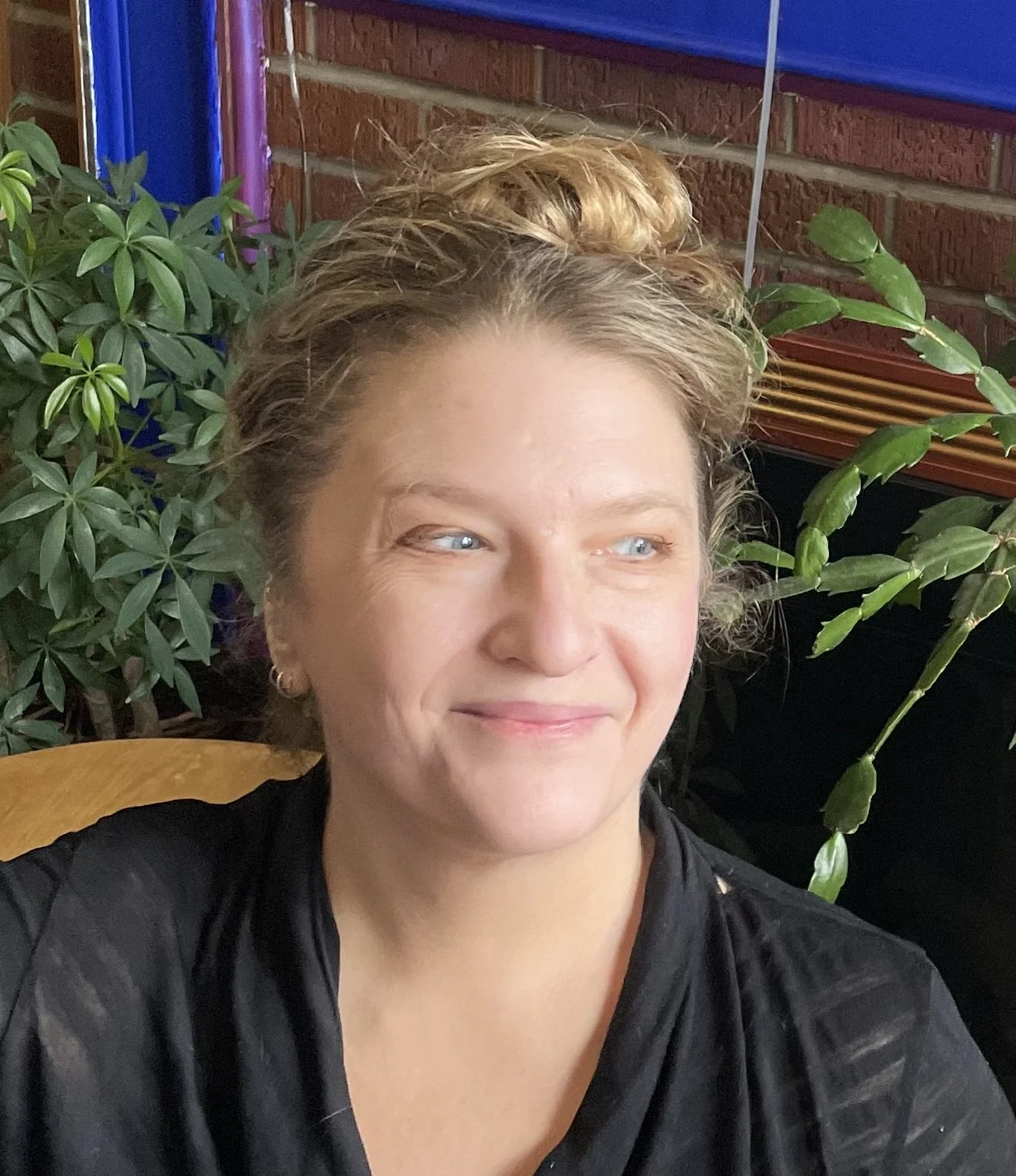 Cecily is a white woman with curly blonde hair escaping from a bun. She is pictured against a background with bricks and plants, smiling towards the side.