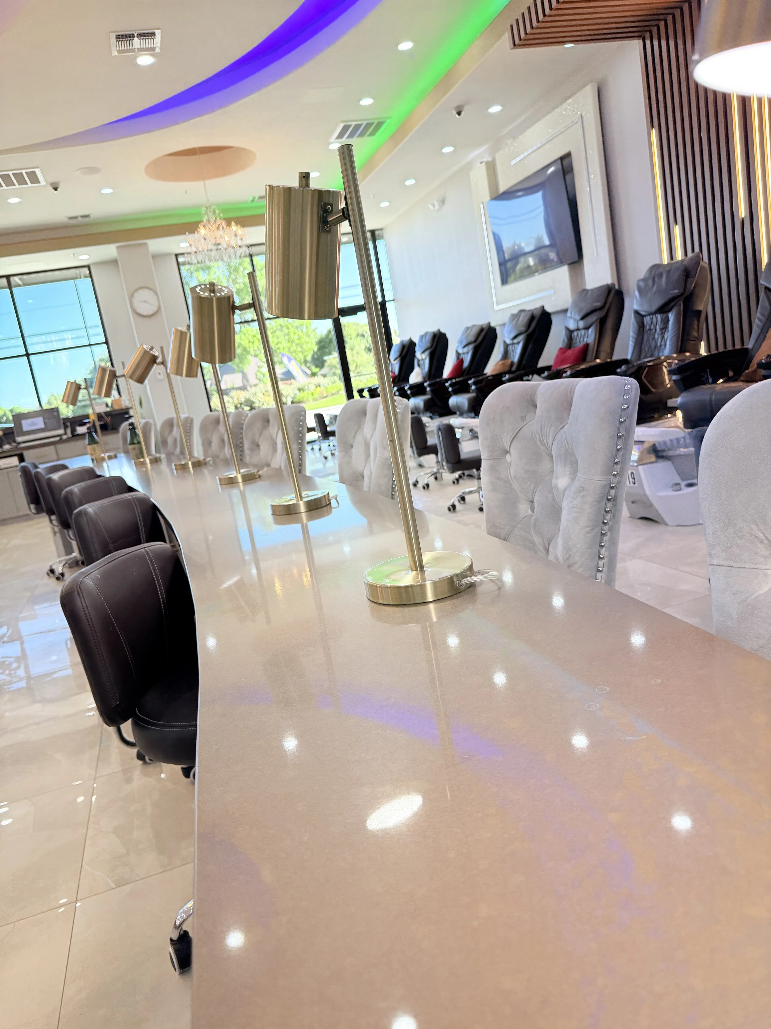 Empty nail salon with pedicure chairs against the wall and a long counter with black and gray chairs in the foreground. Sunlight streams through large windows.
