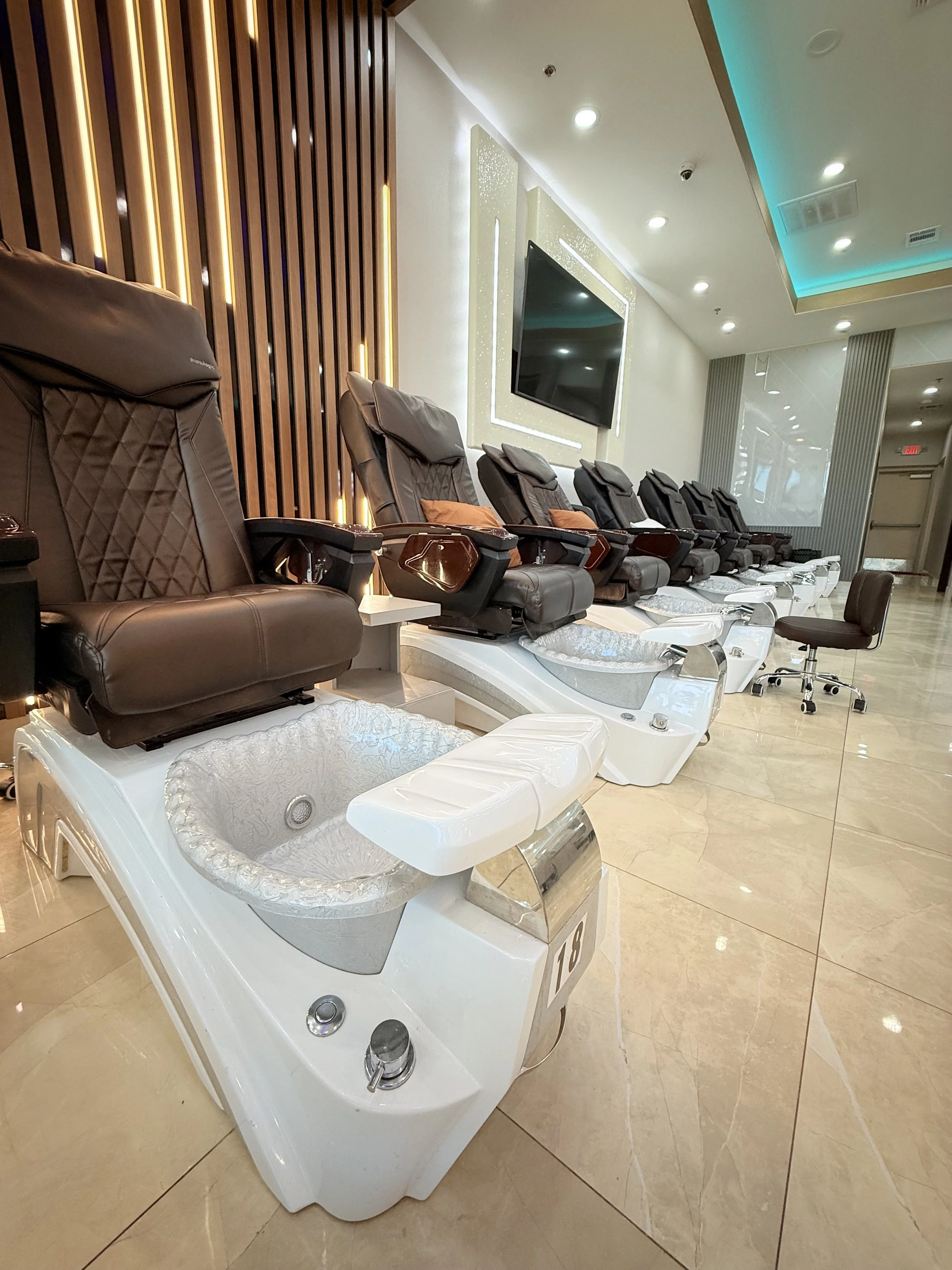 Multiple pedicure chairs with massage features and foot baths in a modern spa or salon, arranged in a row with a television on the wall behind.