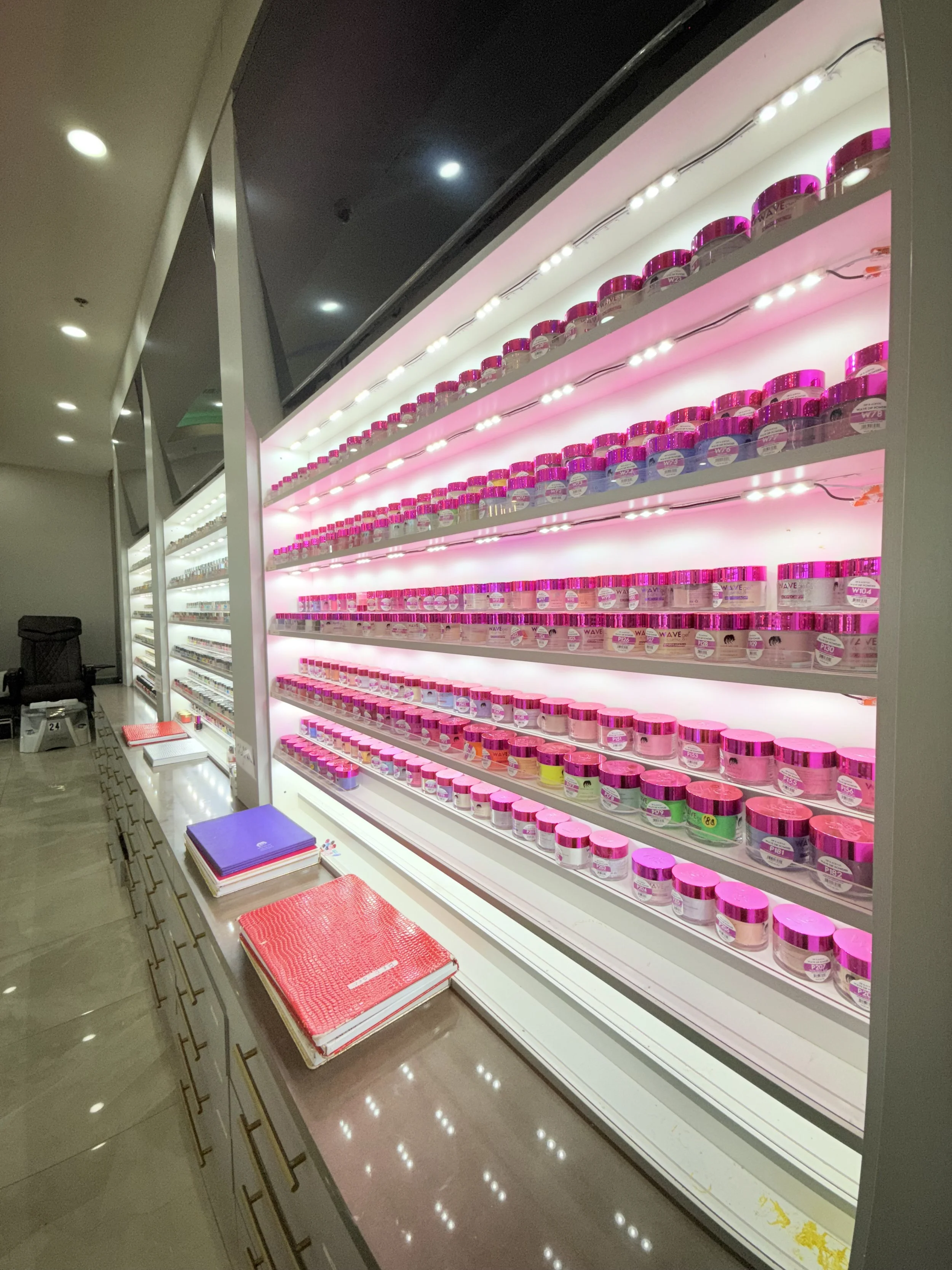 Display of multiple pink-lidded jars with skincare or cosmetic products on white shelves, illuminated by bright pink and white lights, with notebooks and pens on the counter.