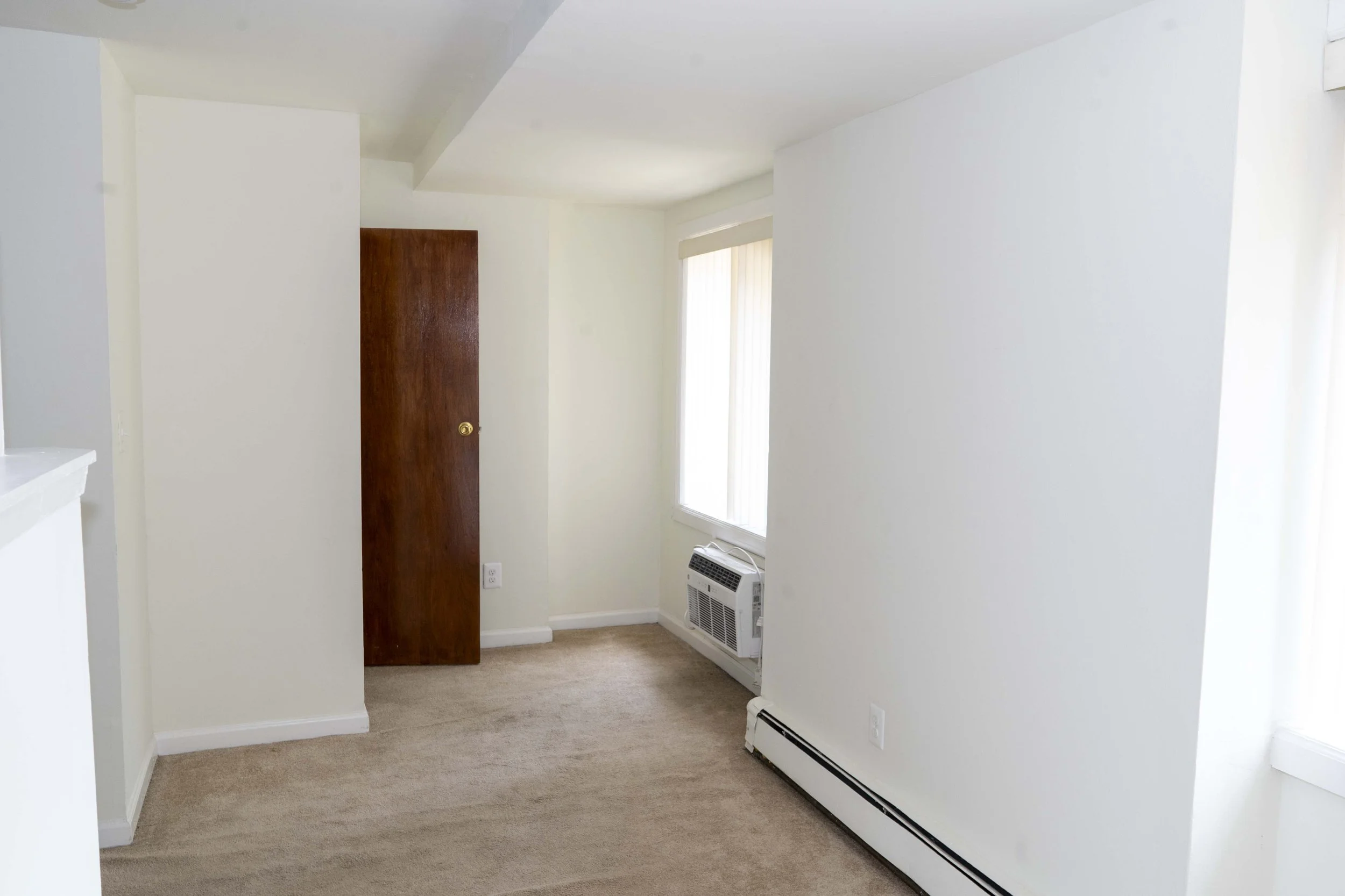 Empty room with beige carpet, white walls, a window with vertical blinds, a wooden door, and an air conditioning unit.