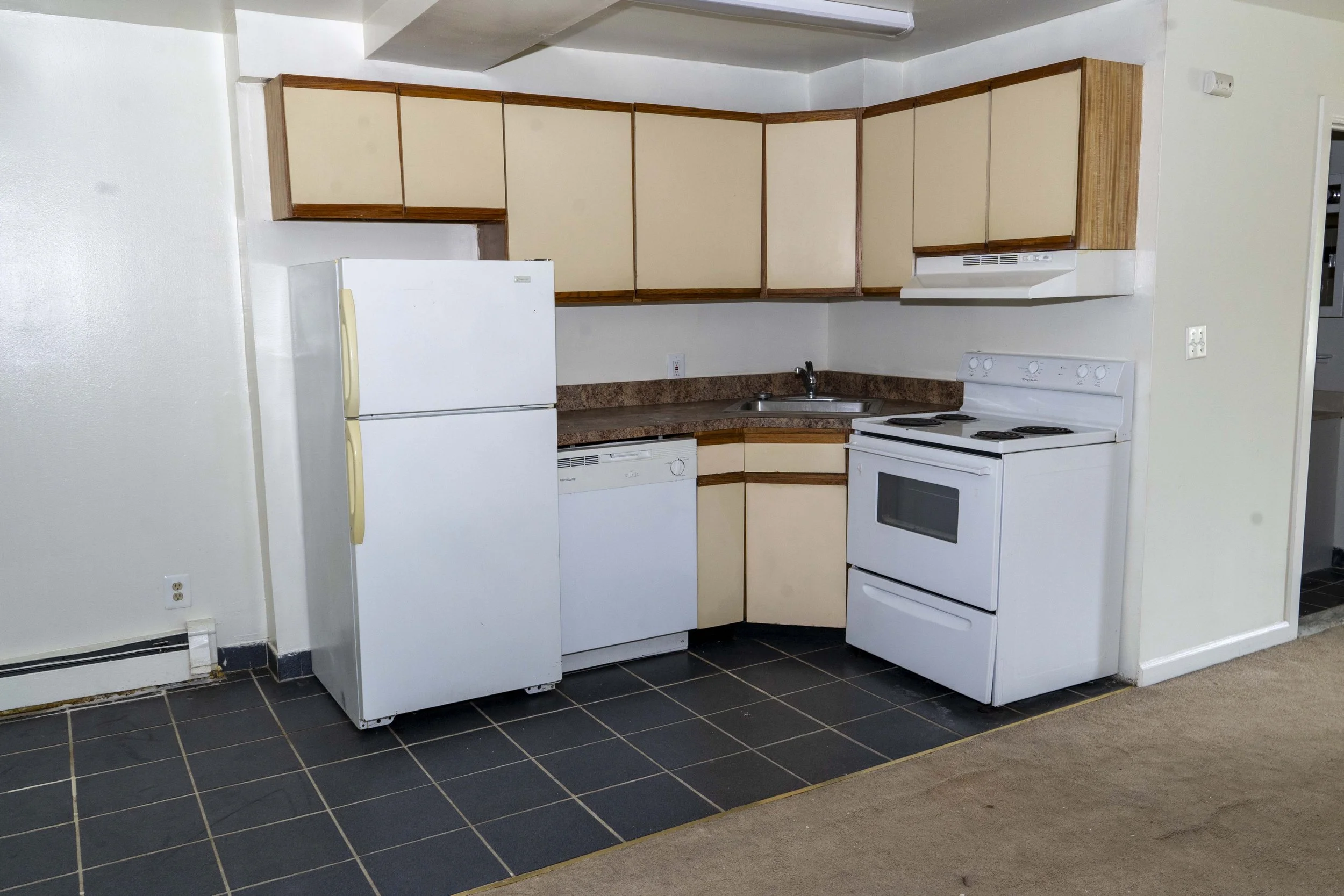 Small kitchen with beige cabinets, white refrigerator, white stove, dishwasher, and a sink. The floor is black tile next to beige carpet.