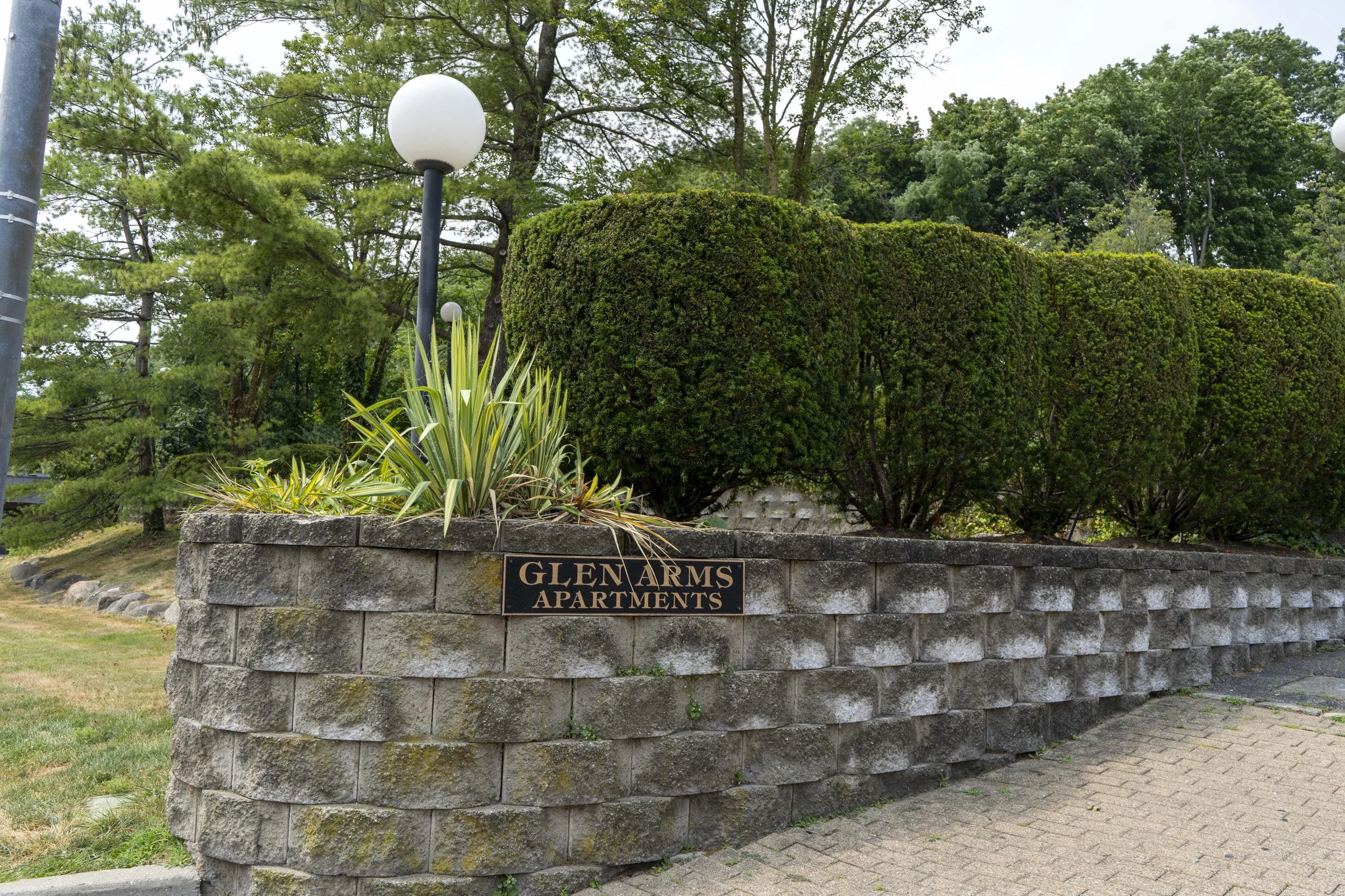 Stone wall with a sign that reads 'Glen Arms Apartments,' greenery including bushes, trees, and lamp posts in the background.