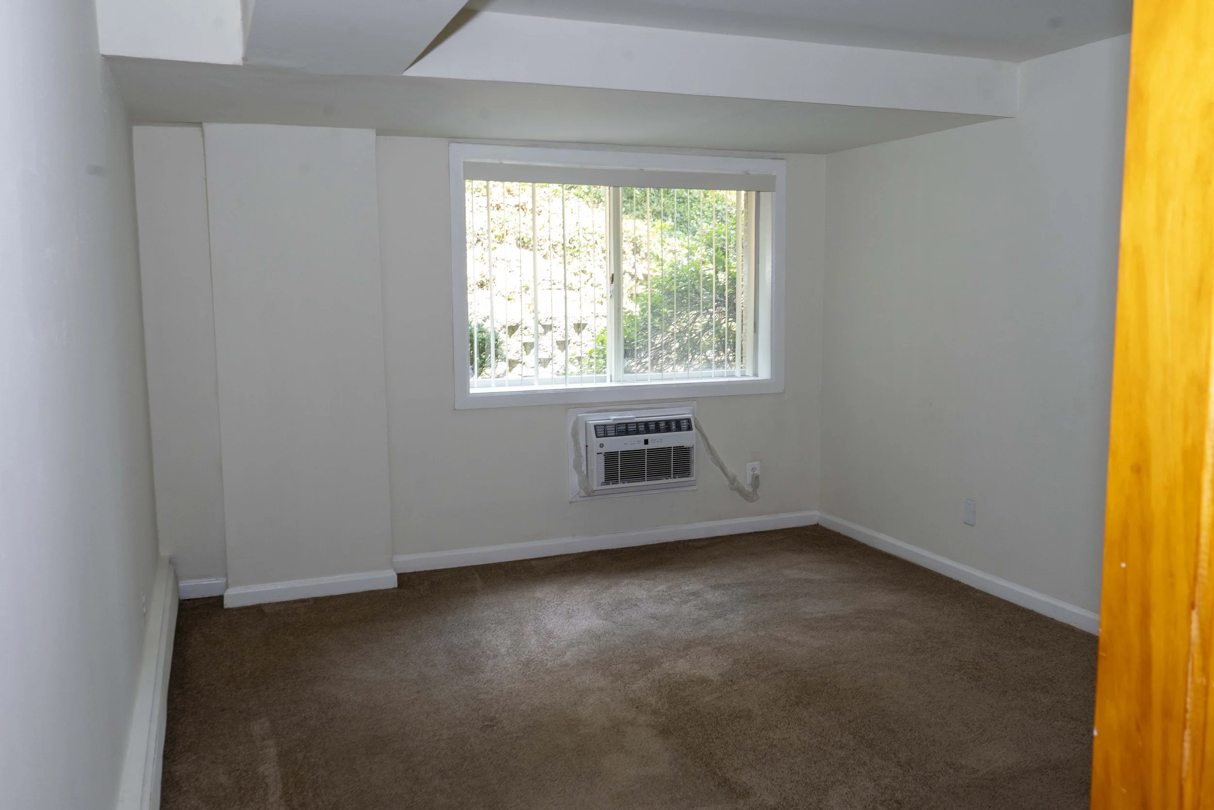 Empty room with a window behind a wall-mounted air conditioner, beige carpet, white walls, and an open door frame on the right side.