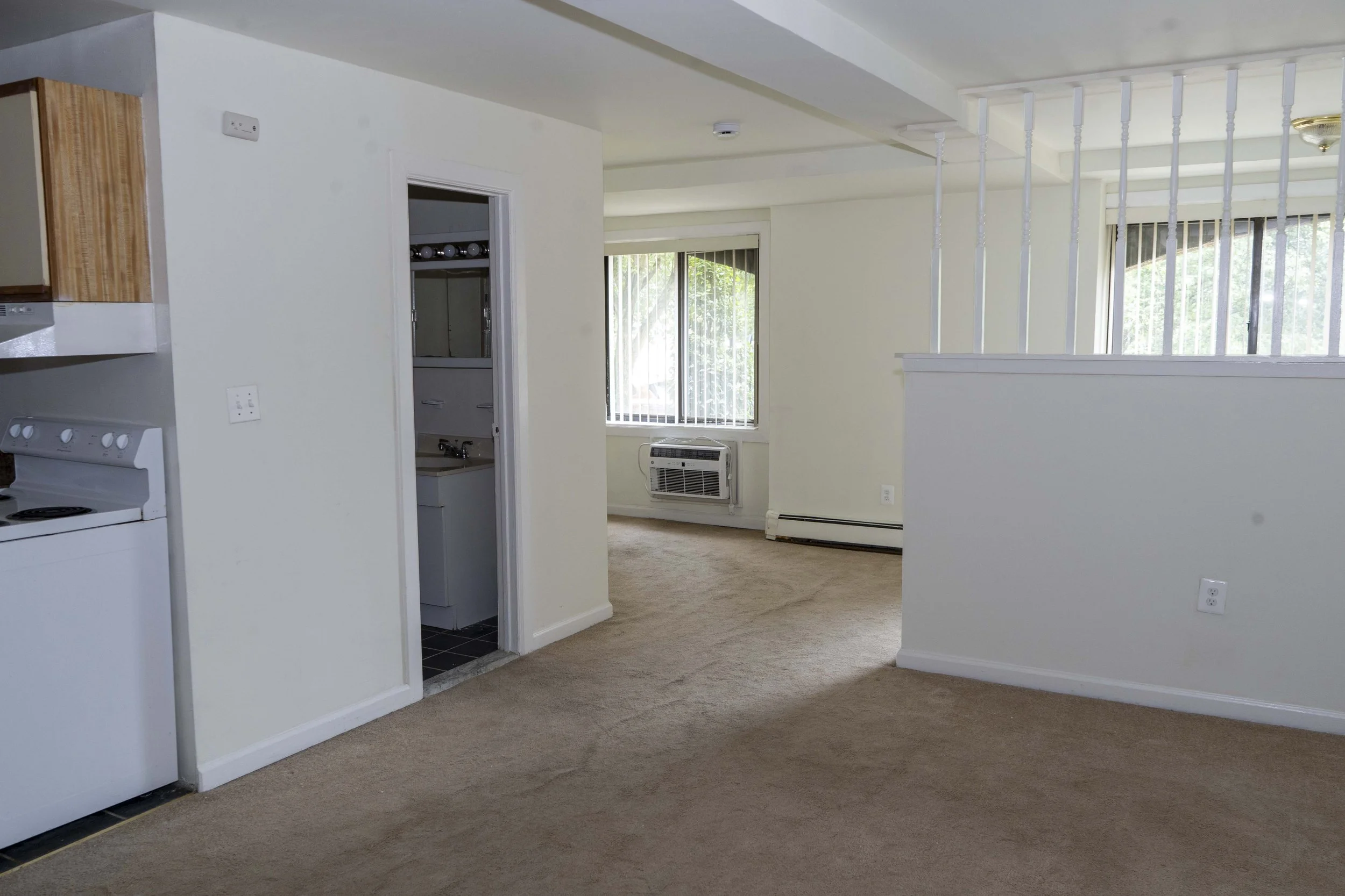 Interior view of an apartment with beige carpet, white walls, a small kitchen area, window air conditioning, and large windows with vertical blinds.