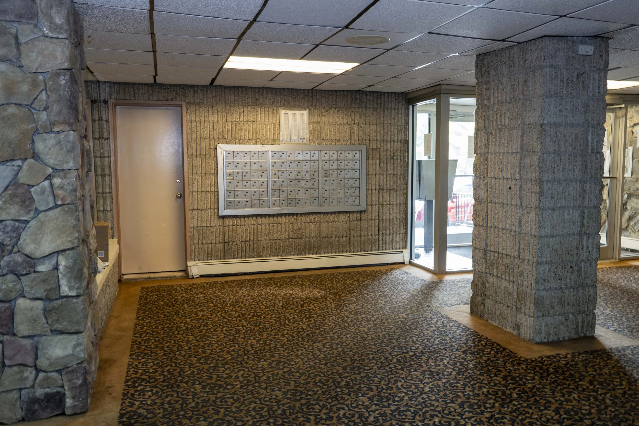 Lobby area with mailboxes on a textured brick wall, a door, and a glass entrance door with natural light.