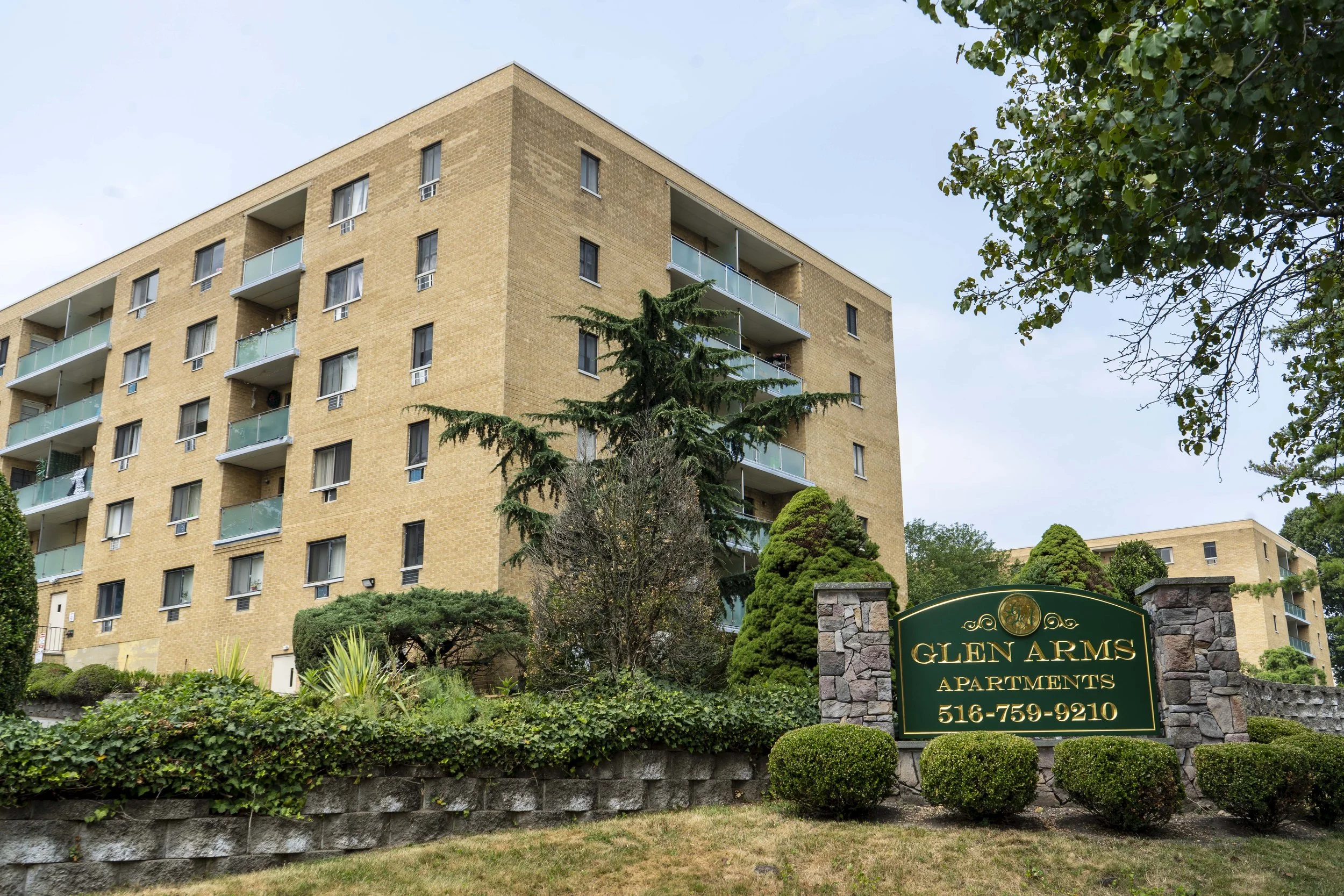 Apartment building with a sign that reads 'Glen Arms Apartments, 516-759-9210' surrounded by landscaped bushes and trees.