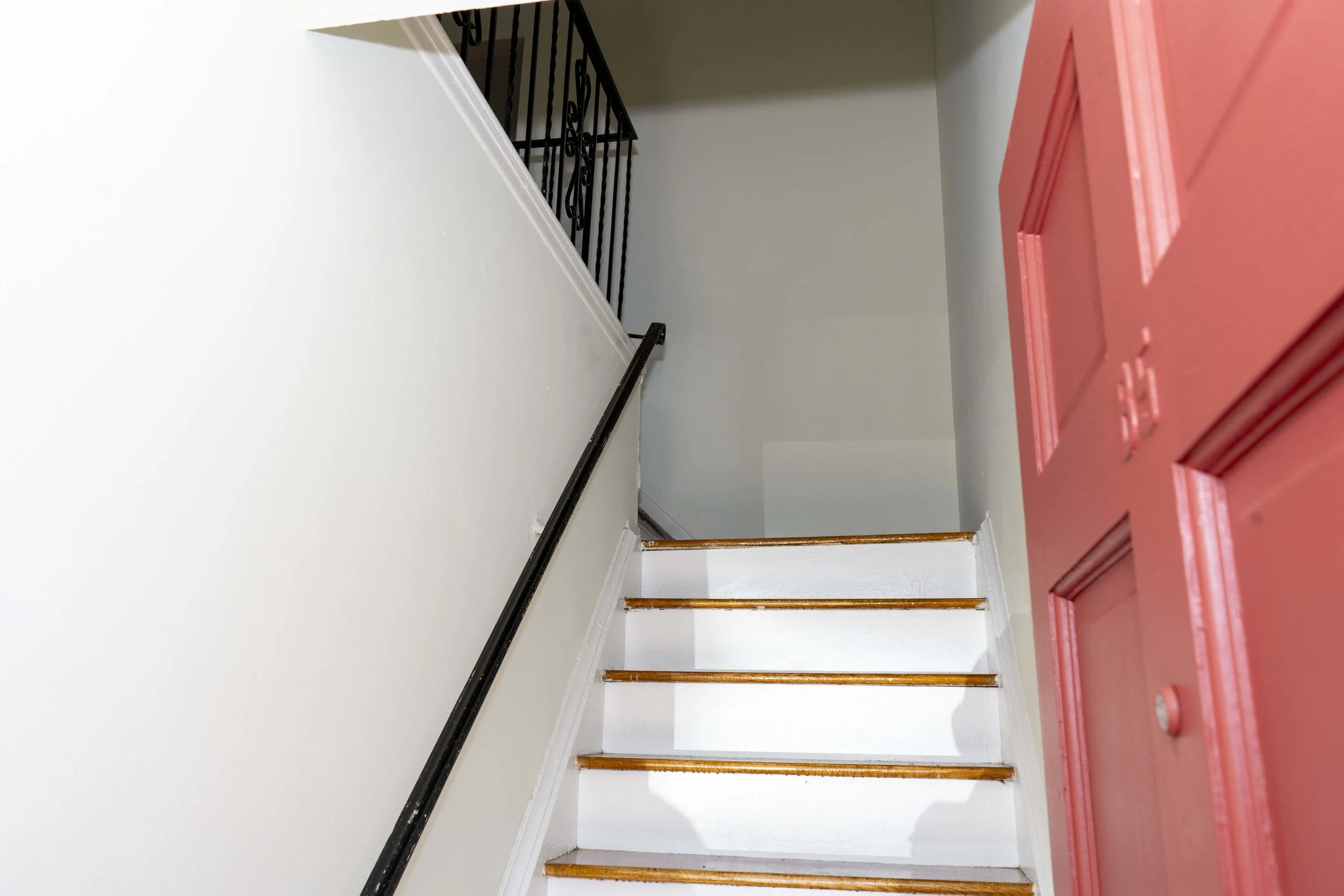 View of a staircase with white risers and wooden treads, black handrail on the left, pink front door on the right, and a black iron railing at the top of the stairs.